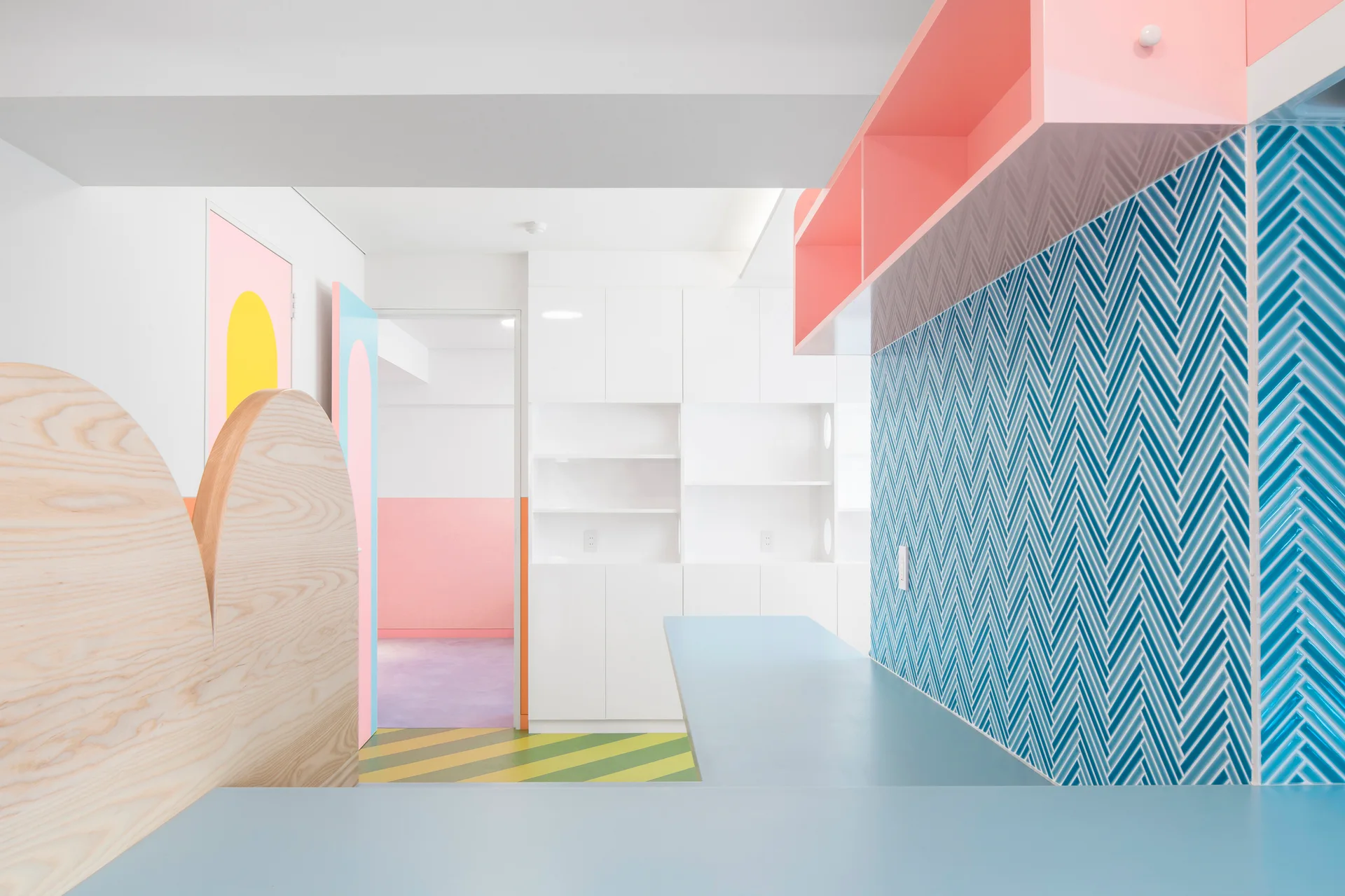 A pastel kitchen features a blue herringbone tiled backsplash, a pink open shelf, a light wood curved divider, and white cabinetry.