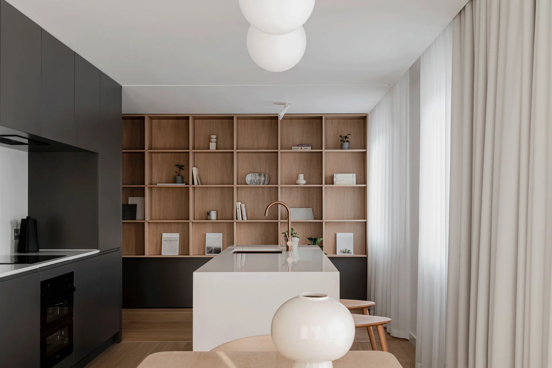 A modern kitchen features a sleek white island with a copper faucet, dark grey cabinetry, and a large grid-style wooden shelving unit.