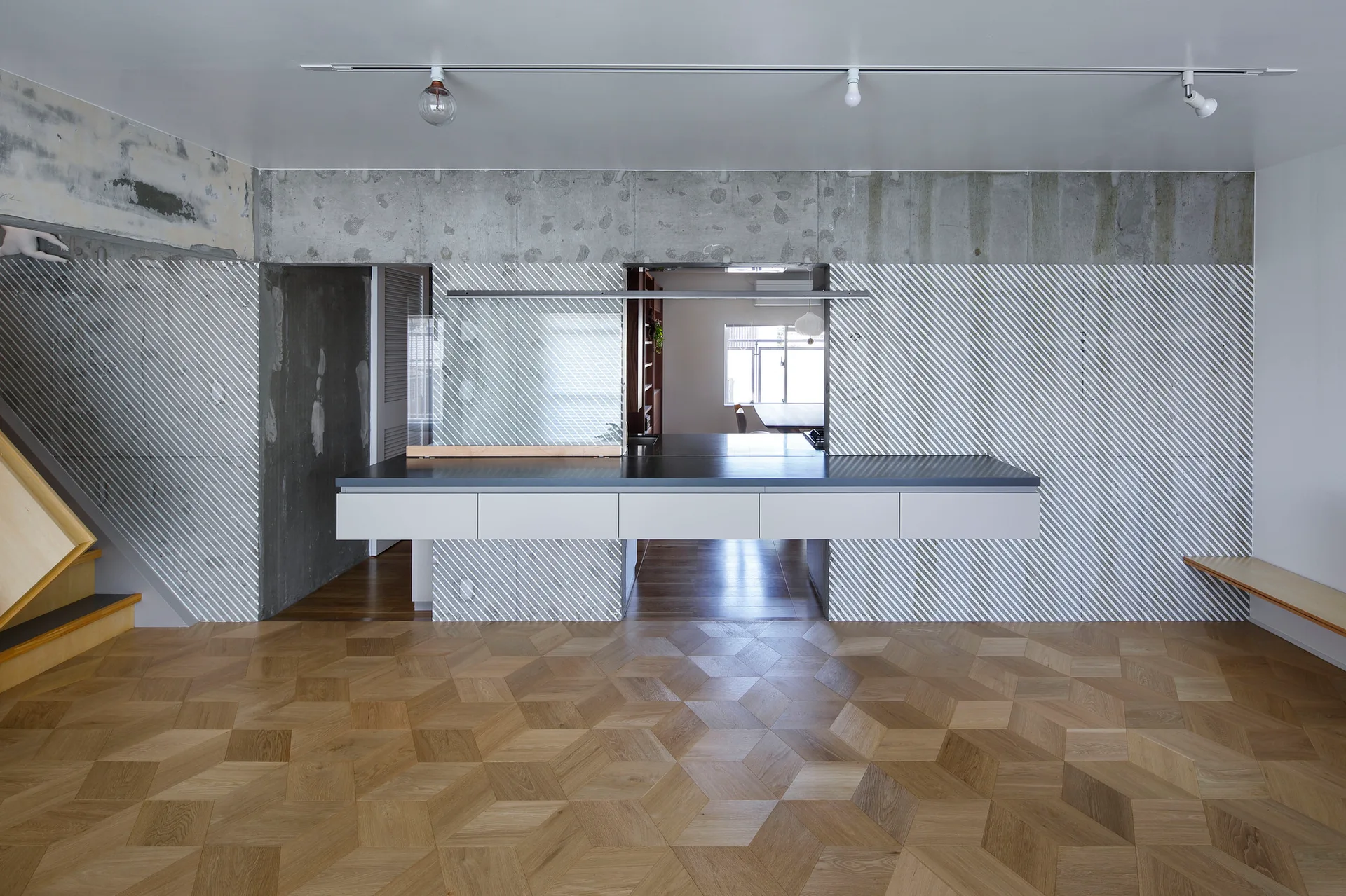 A minimalist kitchen features a long white floating island, diagonal striped wall paneling, an exposed concrete beam, and geometric wood parquet flooring.