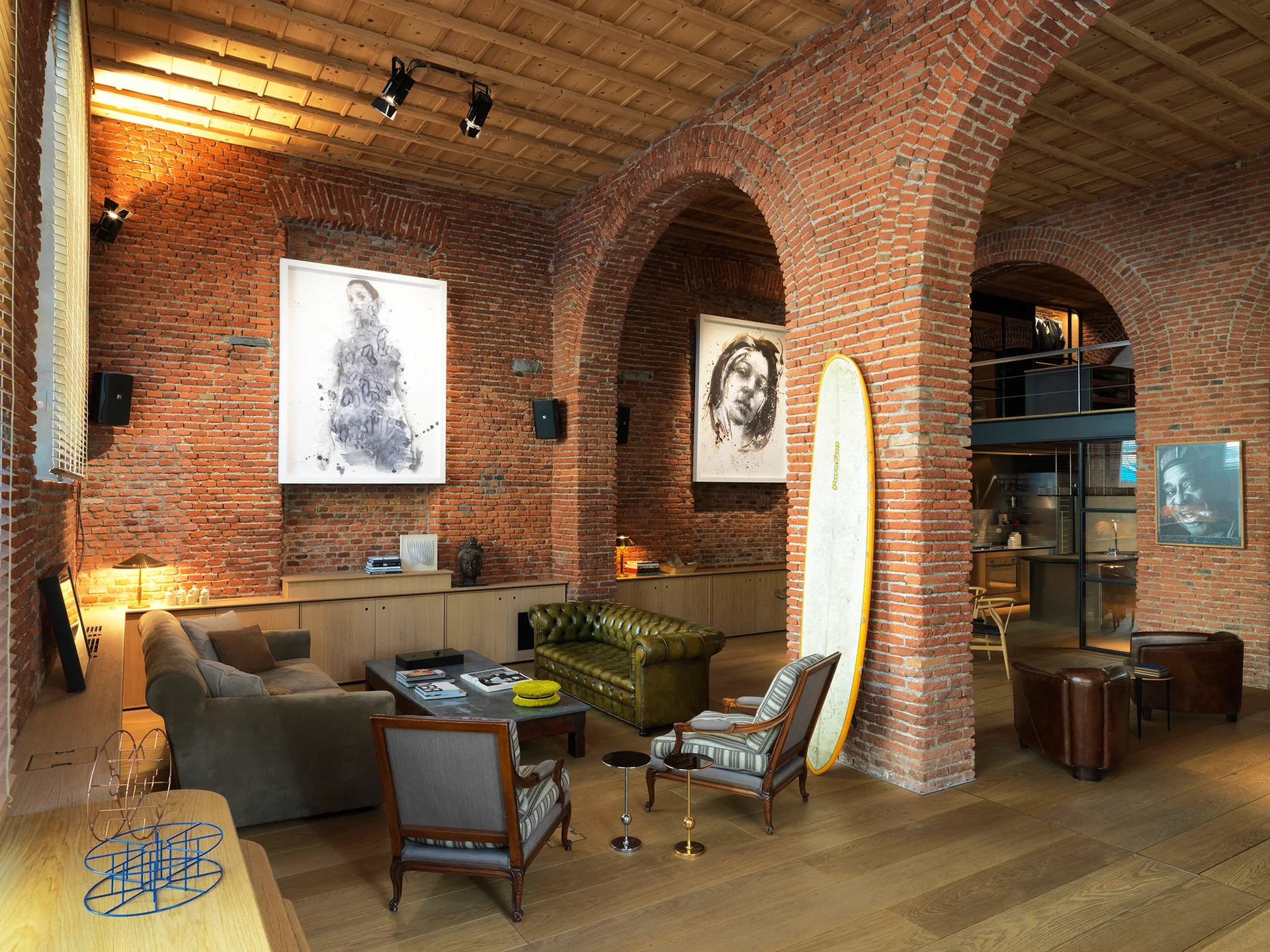 An industrial living room features large red brick archways, a wood-paneled ceiling, eclectic sofas, and framed black-and-white portraits.