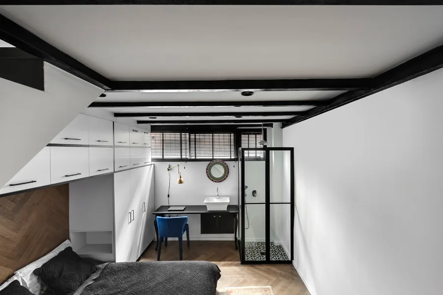 A compact bedroom features a bed with a herringbone wood headboard, white cabinetry, a black desk, and a glass-enclosed shower stall.