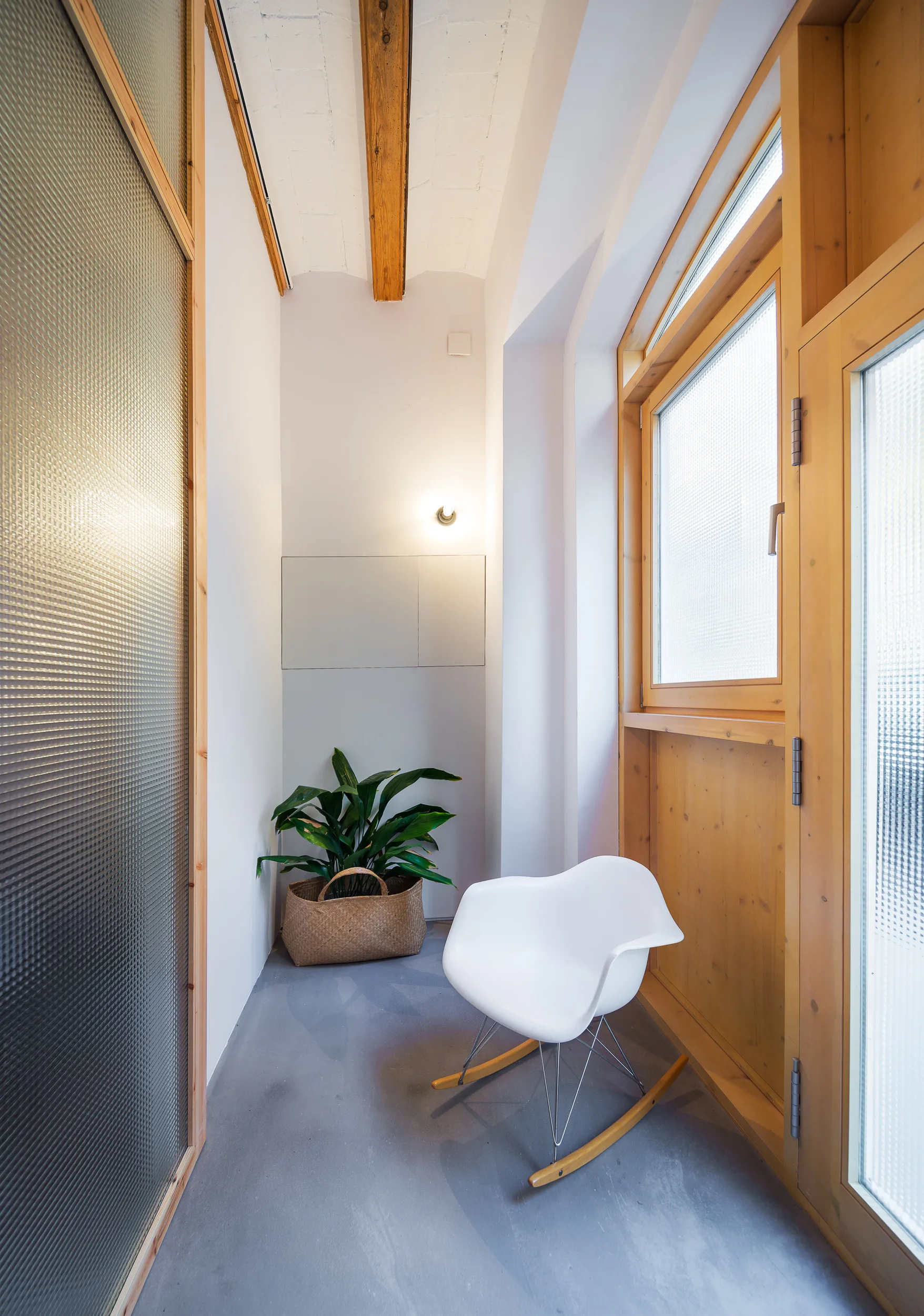 A white rocking chair sits on a grey floor in a hallway next to a wood-framed window and a ribbed glass partition.