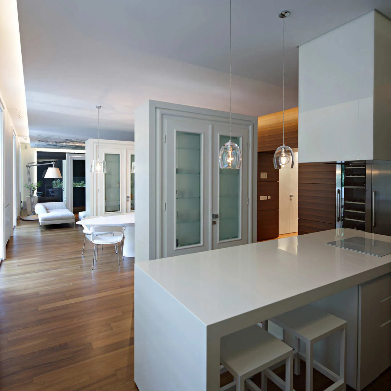 A modern kitchen with a white island and glass pendant lights features white storage partitions separating the dining and living areas.