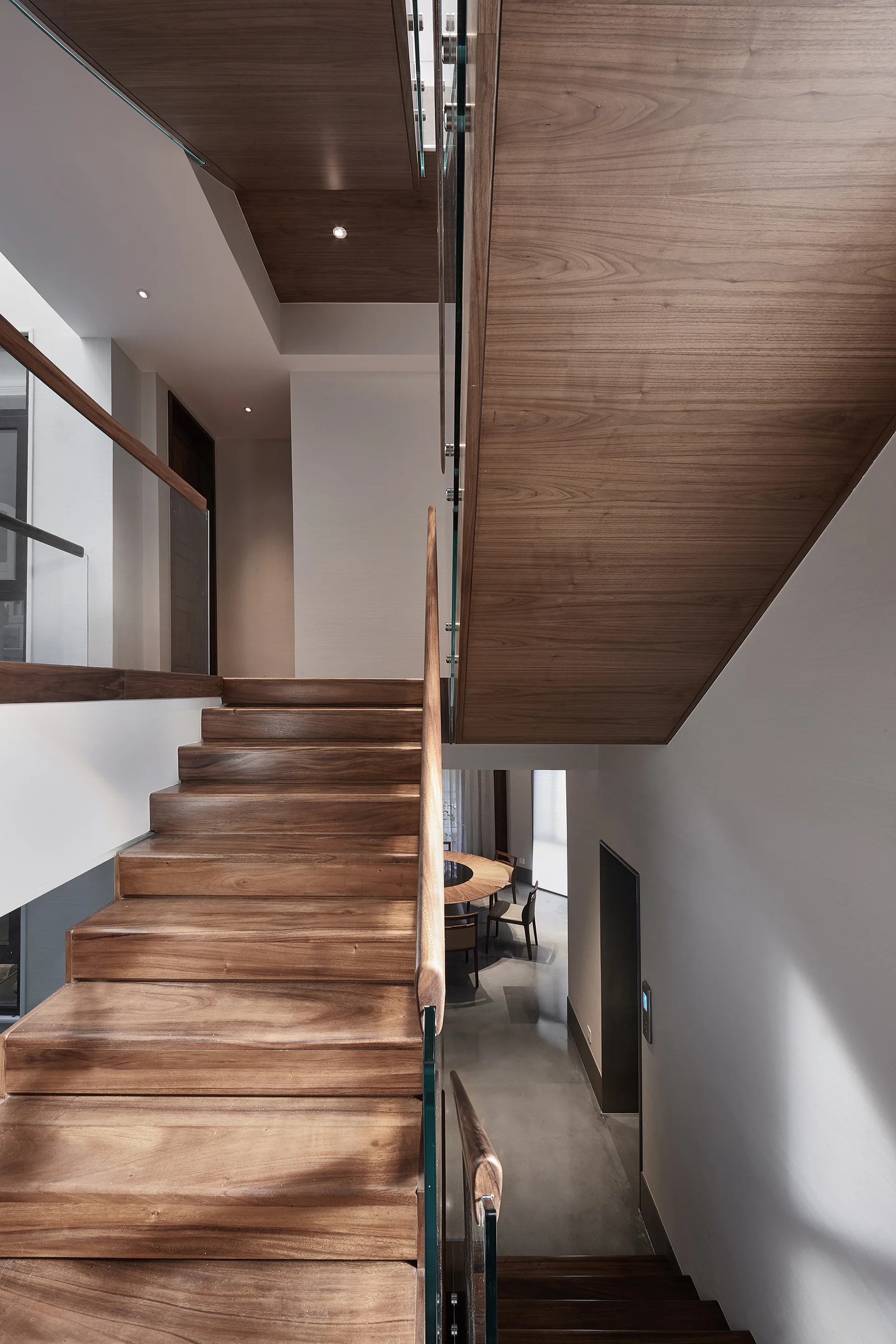 A modern wooden staircase features glass panel railings and overlooks a grey-floored dining space with a round table below.