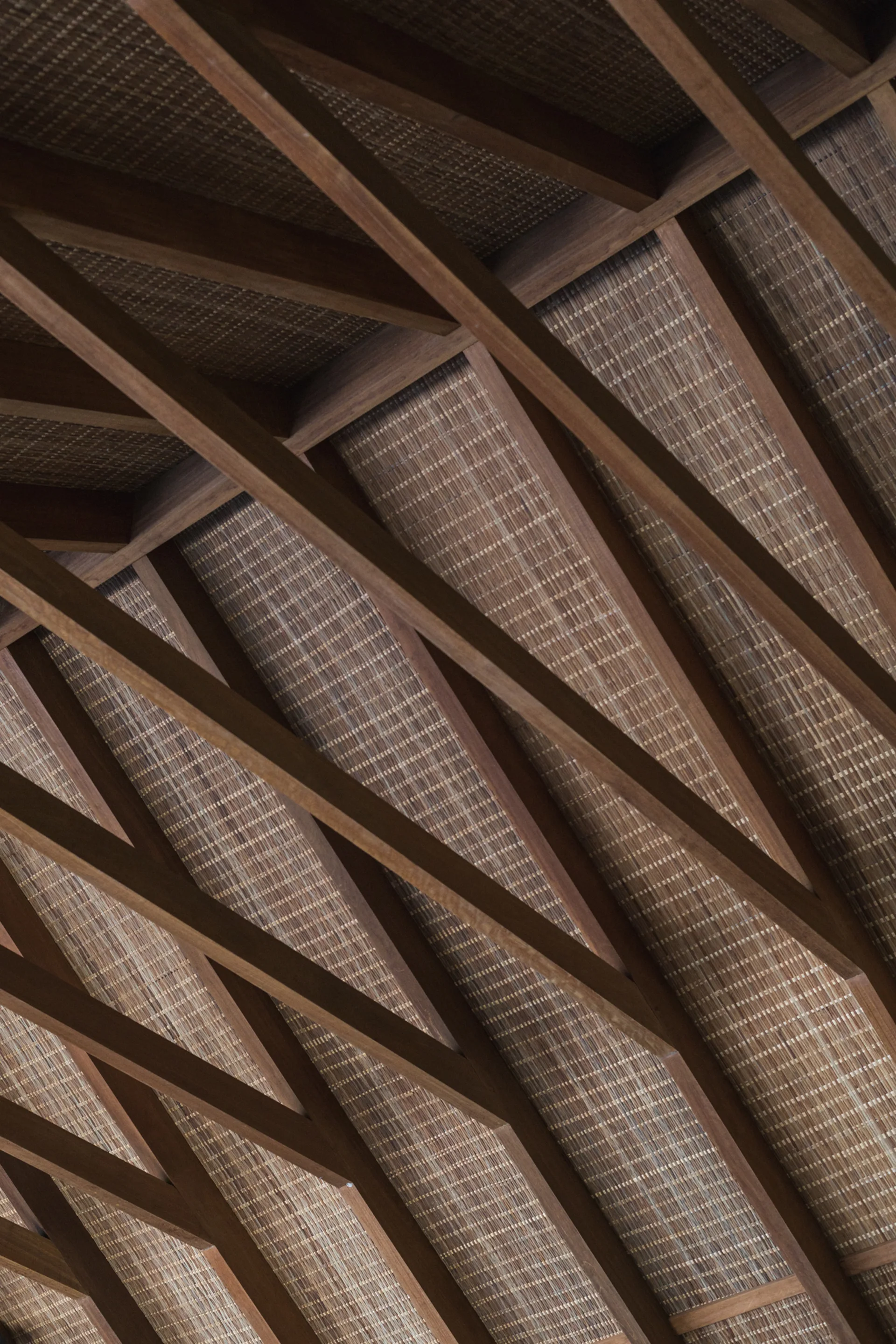 An angled close-up of a ceiling with dark brown wooden rafters layered diagonally over a textured tan woven bamboo backing.