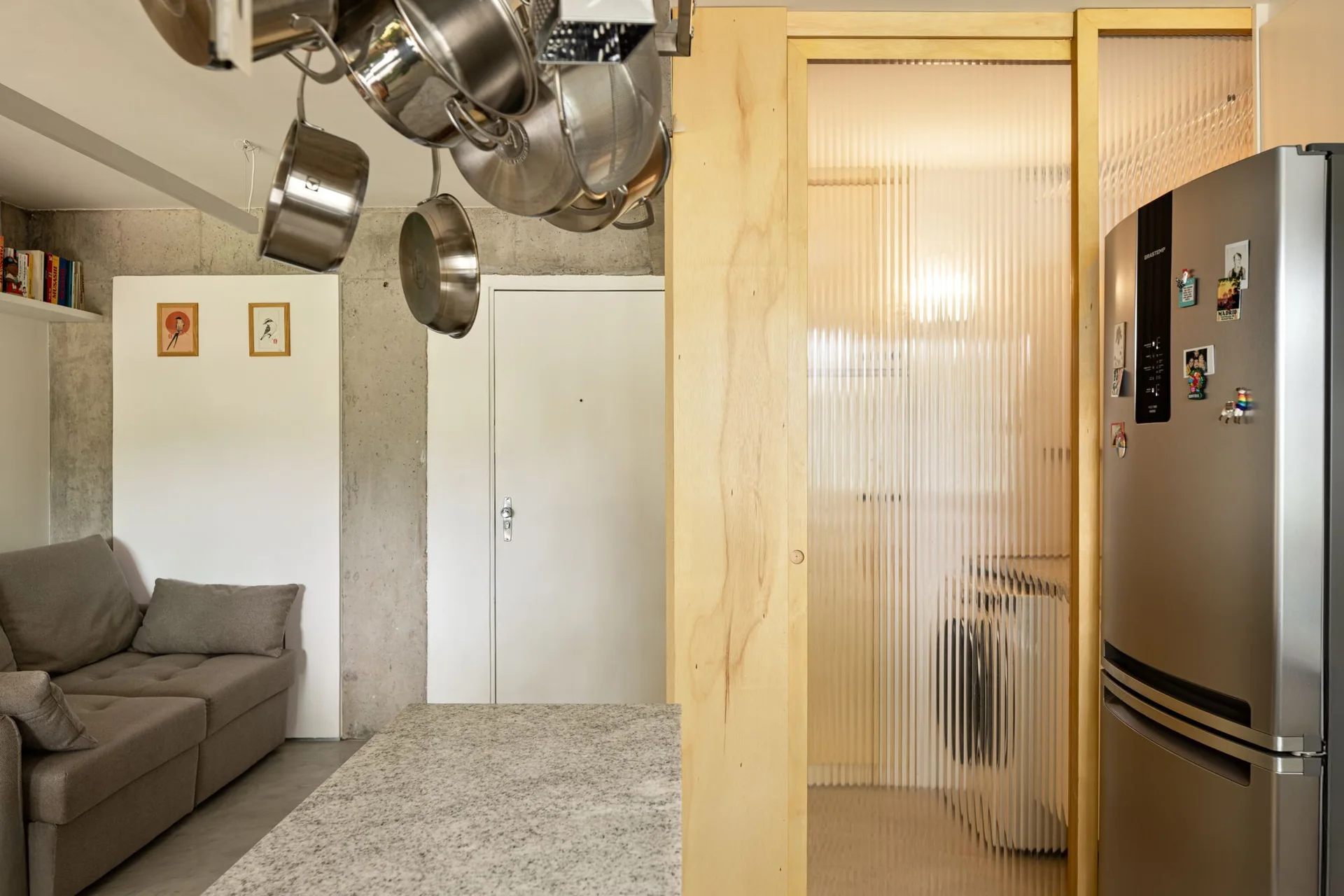 A modern interior features a grey sofa, an exposed concrete wall, a stainless steel refrigerator, and a fluted glass sliding door.