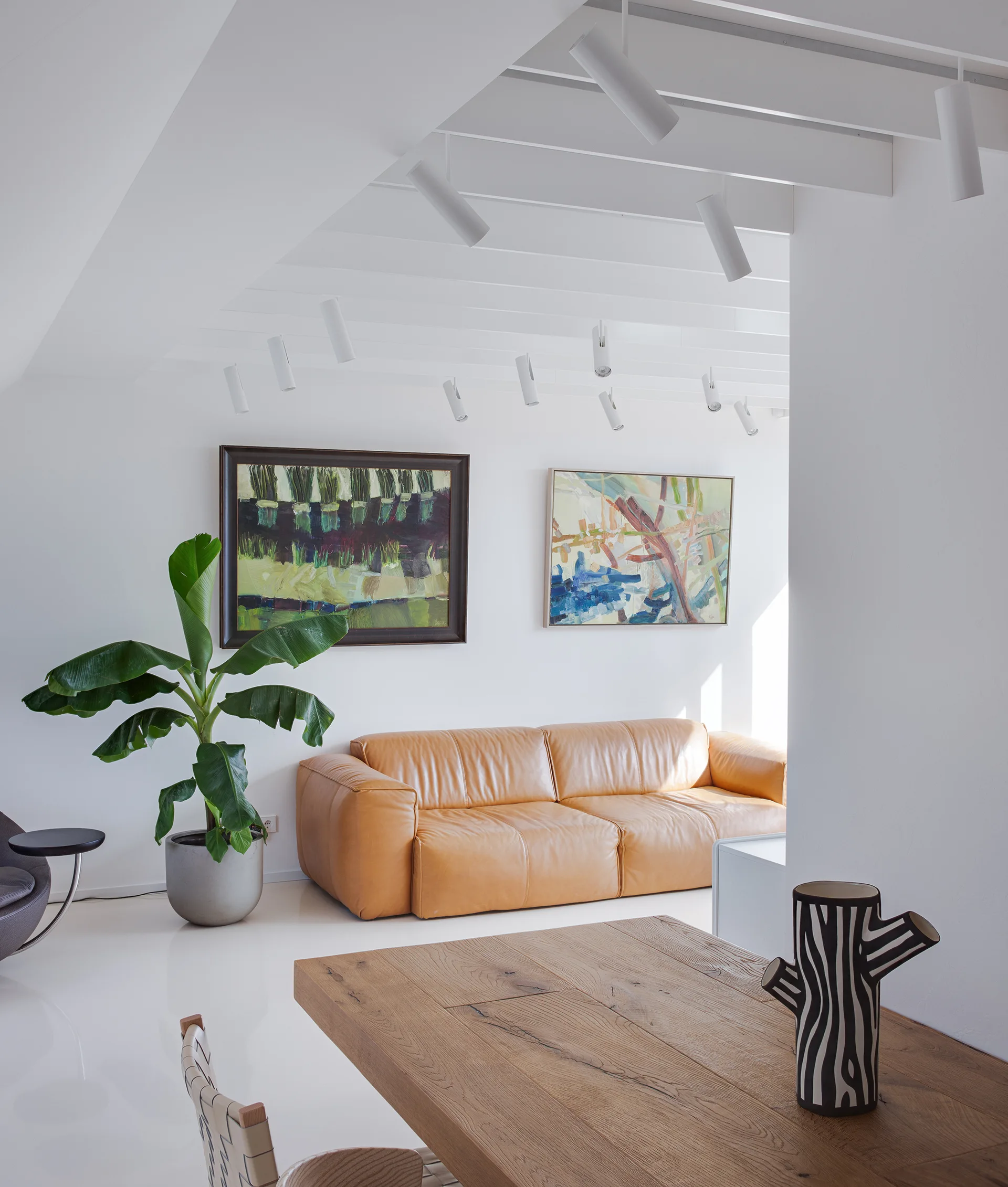 A bright minimalist living space features a tan leather sofa, two abstract paintings, white exposed ceiling beams with track lights, and a wooden table.