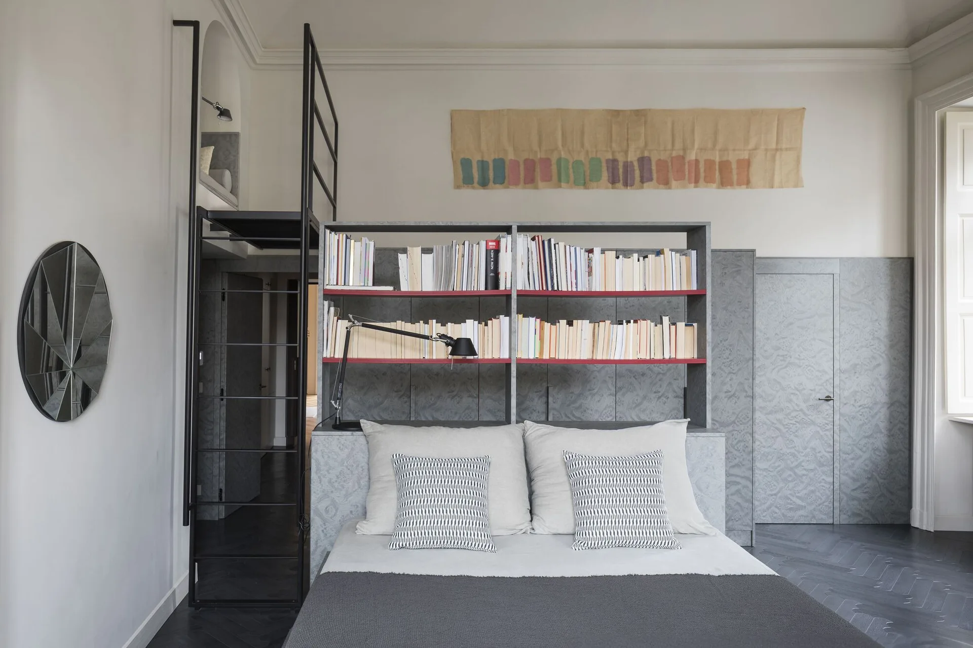 A modern grey bedroom with a bed positioned beneath a built-in bookshelf, next to a black metal ladder and textured wall panels.