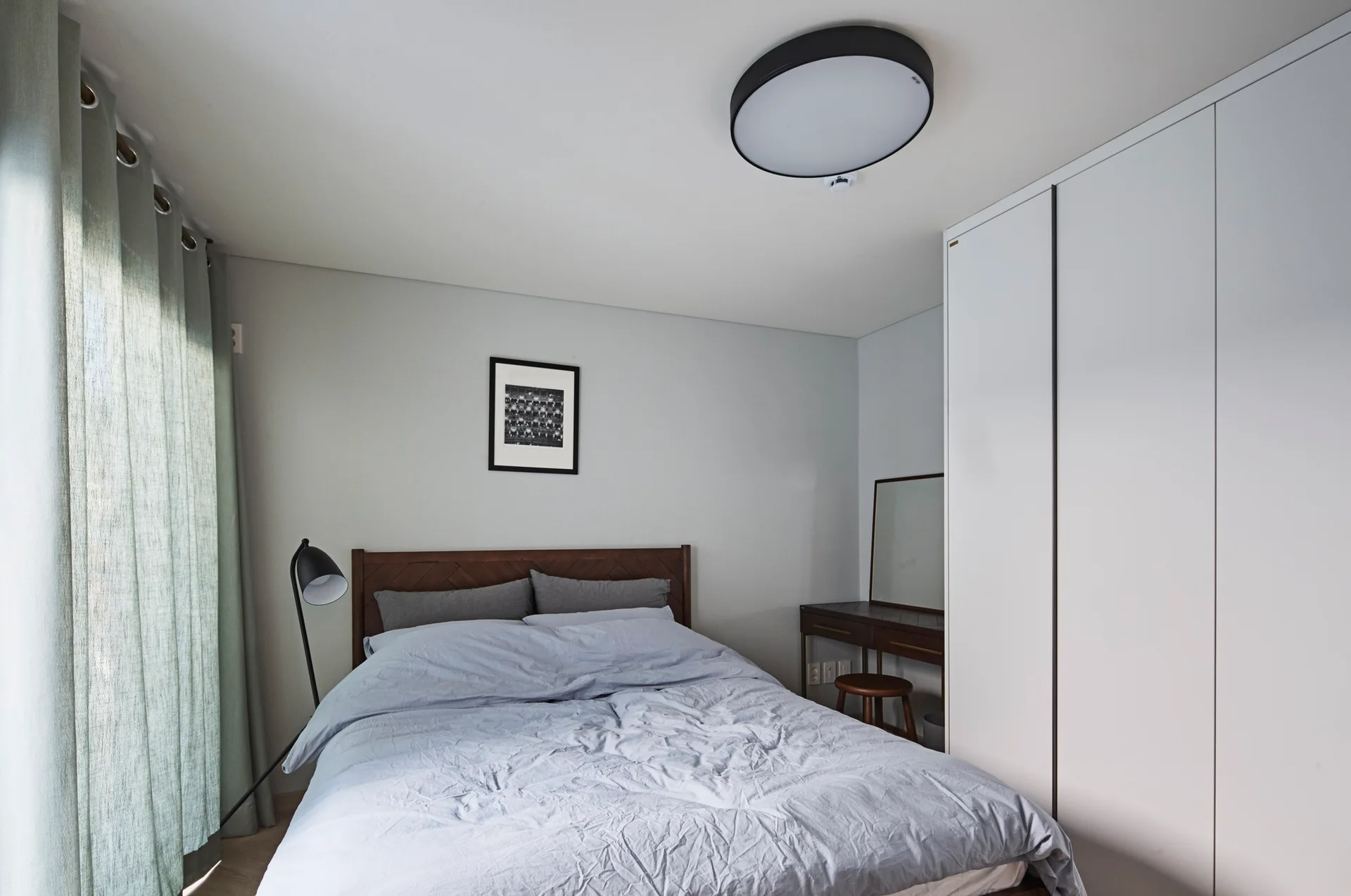 A minimalist bedroom featuring a wooden bed with light blue-grey bedding, a built-in white wardrobe, and light green grommet curtains.