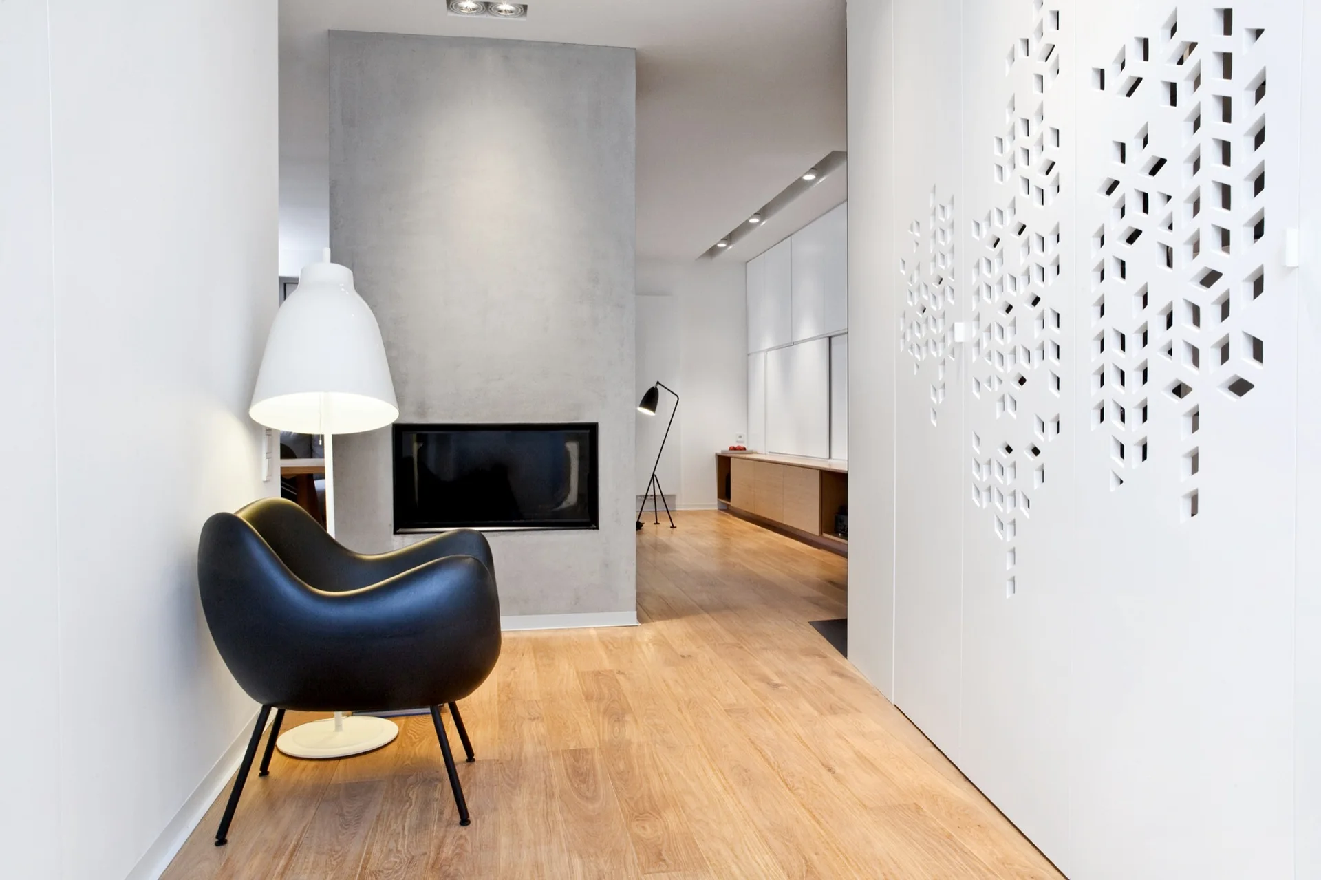 A modern living space featuring light wood floors, a grey concrete fireplace, a black curved armchair, and a white partition wall with geometric cutouts.