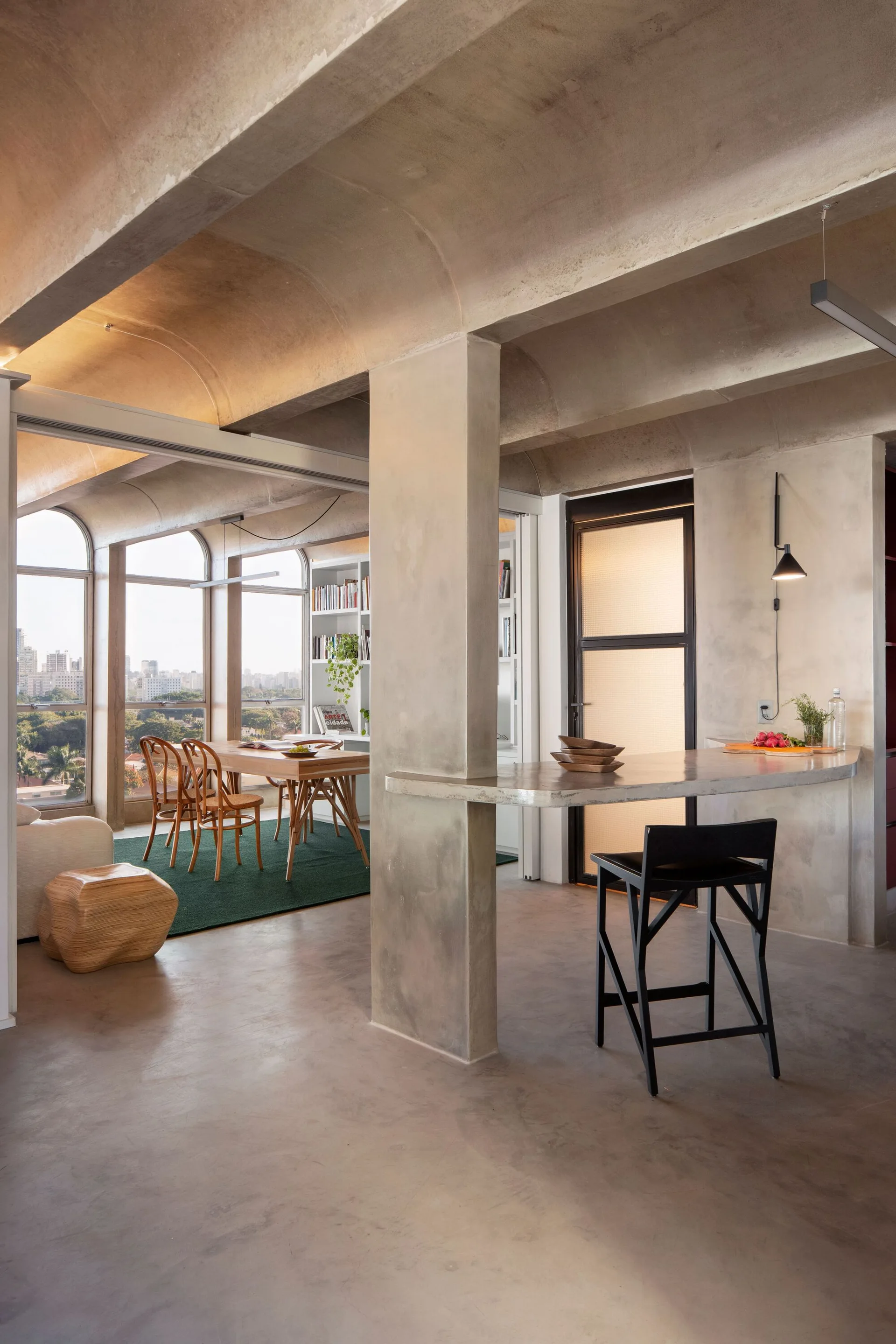 An open-plan room features vaulted concrete ceilings, a custom concrete bar, and a wooden dining table near large arched windows.