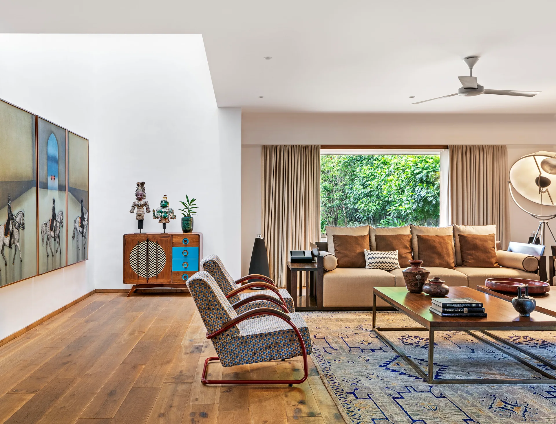 An eclectic living room features a tan sofa, two patterned armchairs, a wooden coffee table, and a large triptych painting on a white wall.
