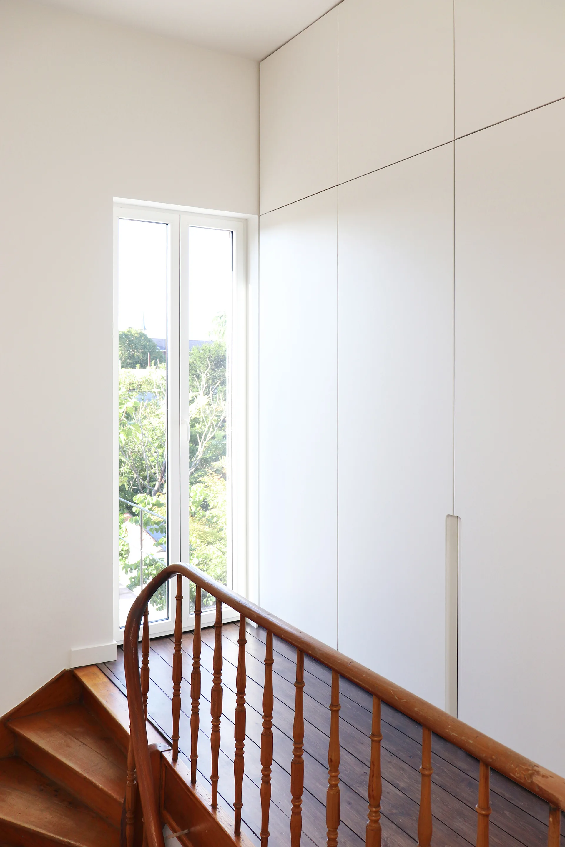 A white staircase landing features a wooden handrail, a tall narrow window, and seamless floor-to-ceiling white storage cabinets.