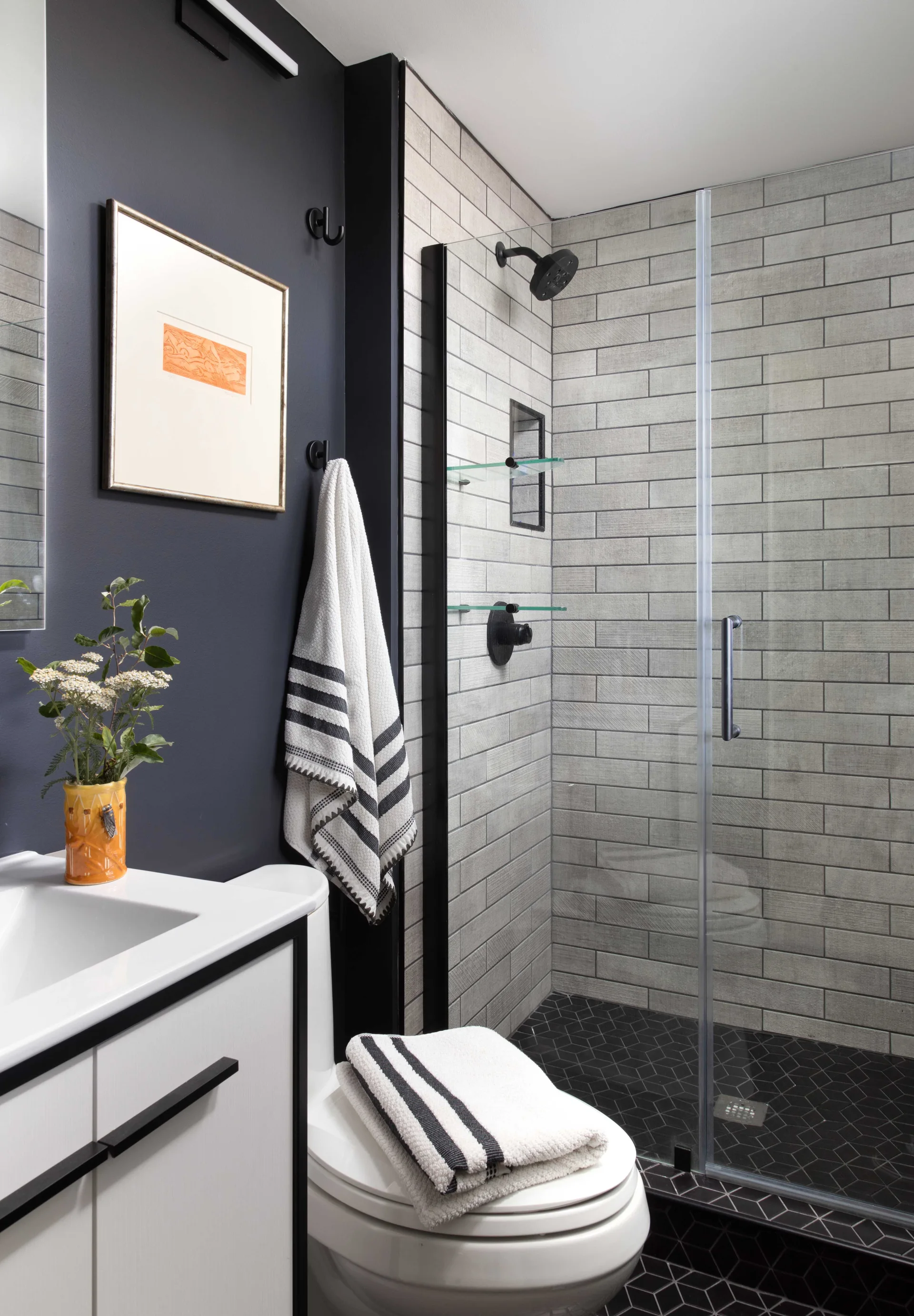 A modern bathroom features a dark grey painted wall, a white vanity cabinet, a toilet, and a glass-enclosed shower with grey rectangular tiling.
