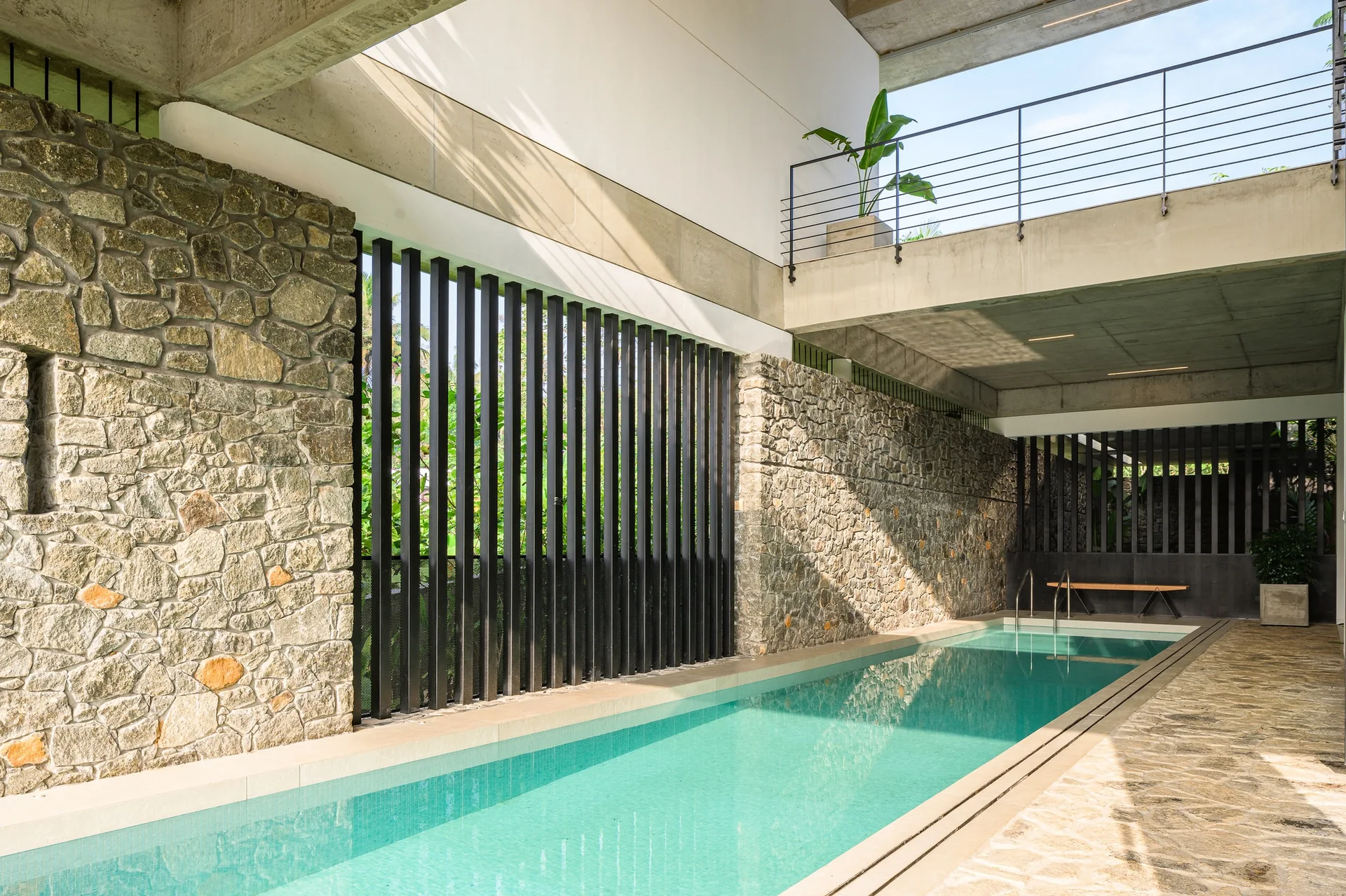 A long rectangular swimming pool flanked by textured rubble stone walls and black vertical slats under an exposed concrete mezzanine.