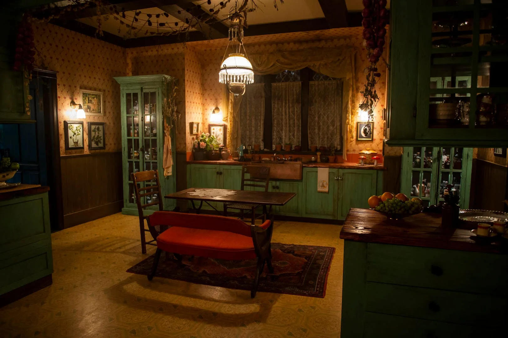 A rustic kitchen featuring distressed teal cabinets, a central wooden dining table with a red bench, floral wallpaper, and warm hanging pendant lights.