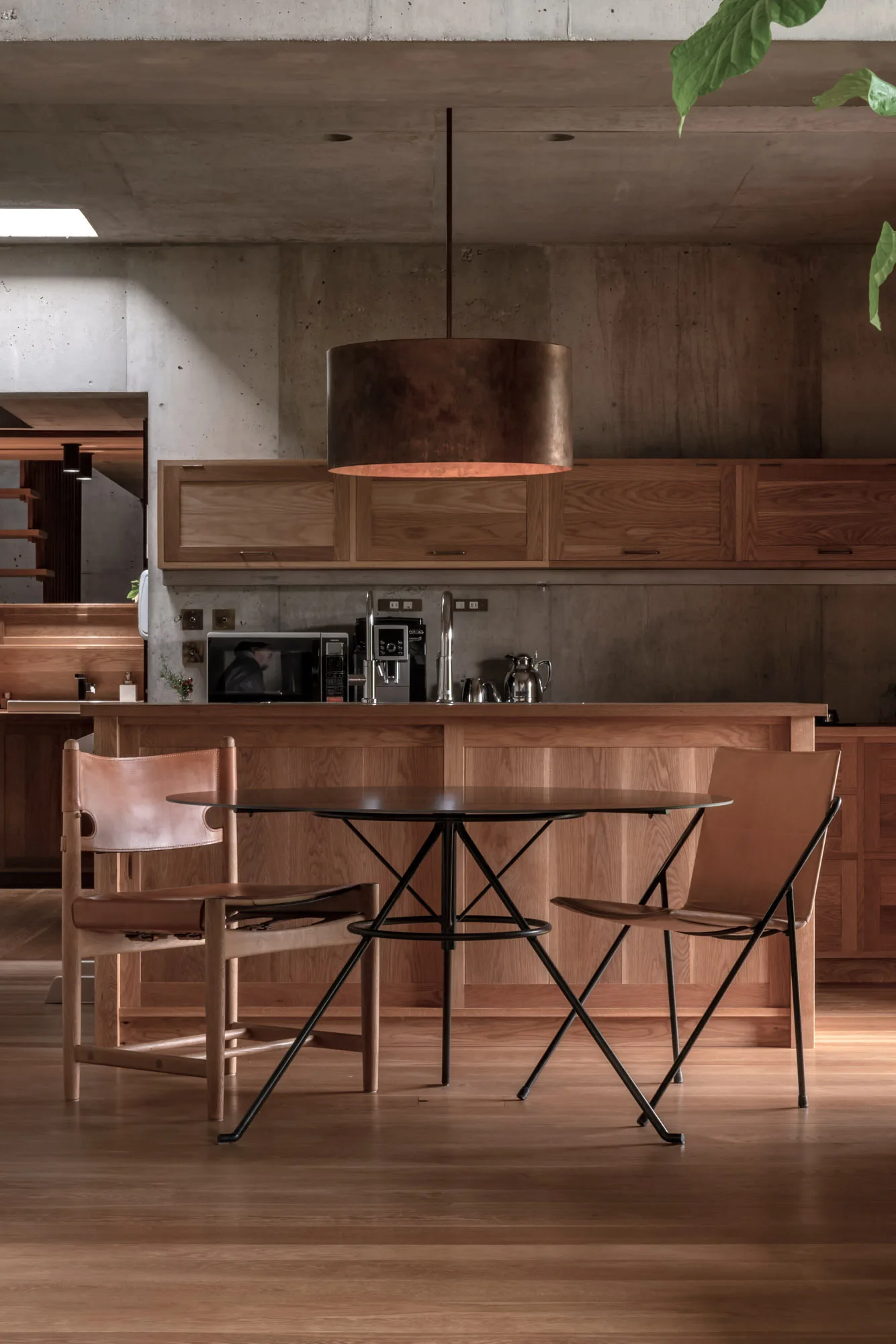 A modern kitchen featuring a wooden island and cabinets against concrete walls, with a round glass dining table and leather chairs in the foreground.