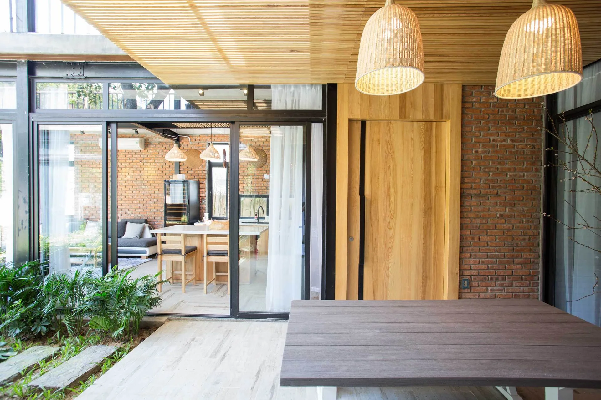 Large black-framed sliding glass doors separate a stone patio from an indoor kitchen, featuring exposed brick walls and a wooden slat ceiling.