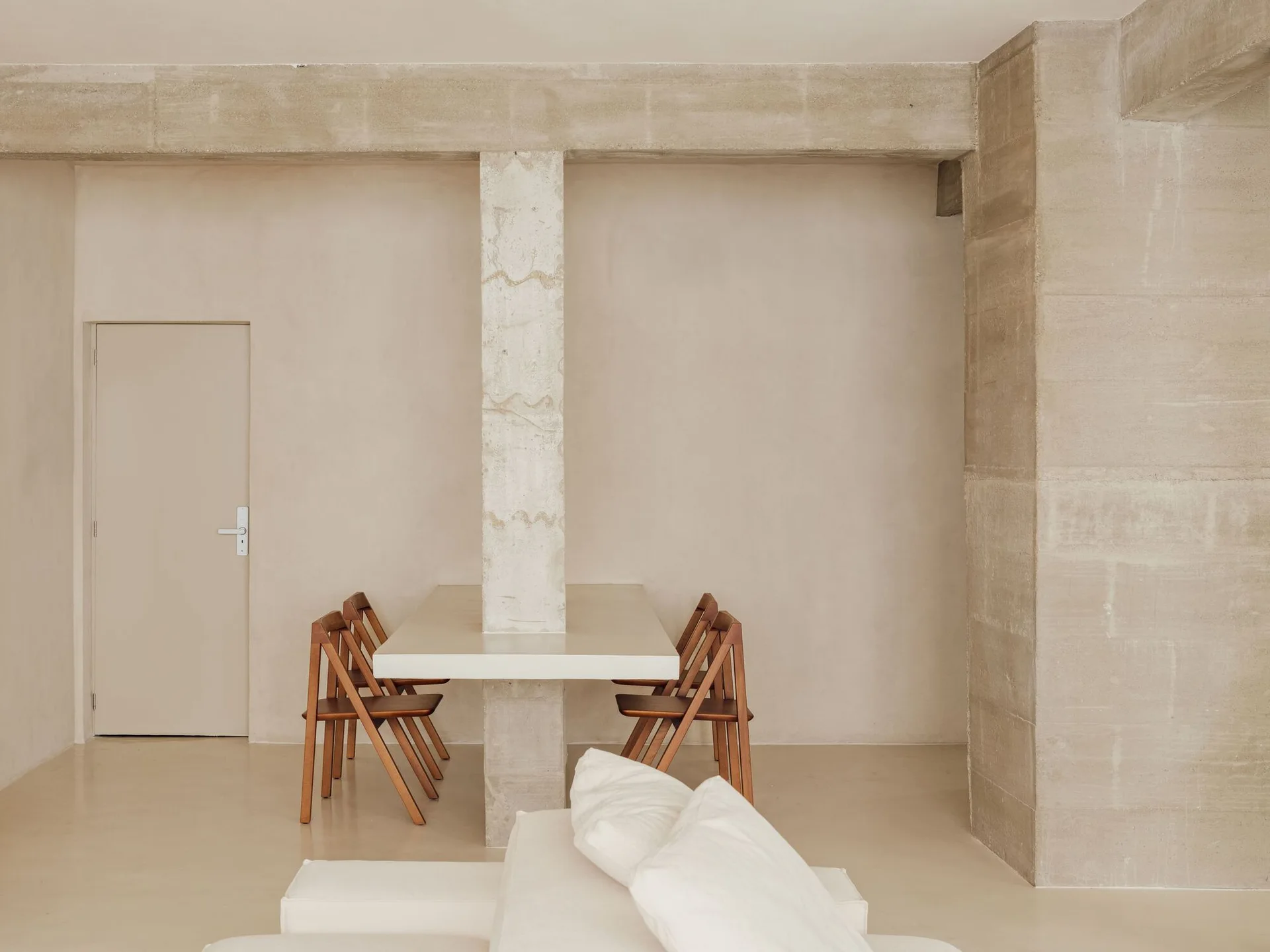 A minimalist dining room featuring a beige table built around a central concrete pillar with four wooden folding chairs.