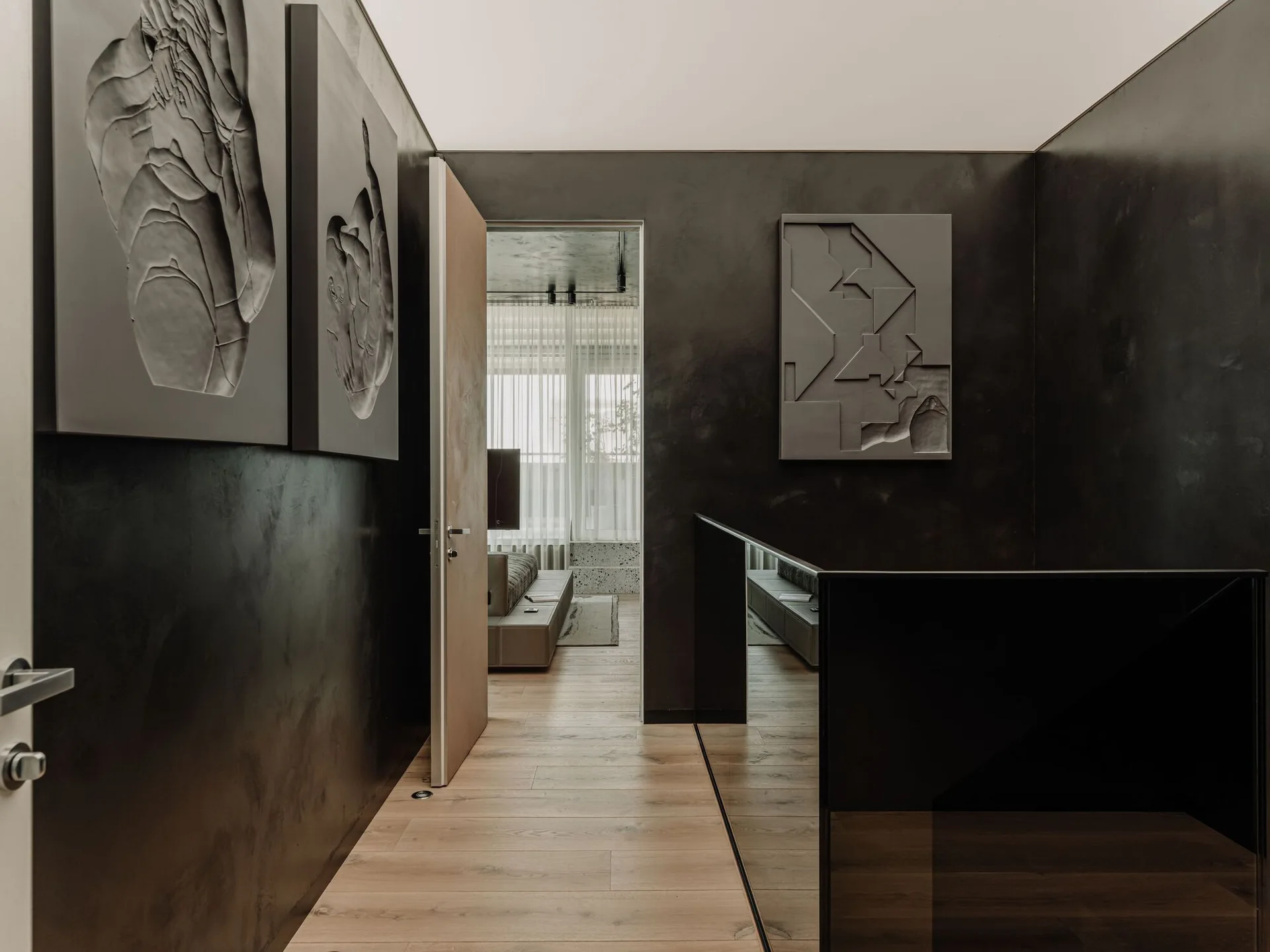 A dark charcoal hallway featuring sculptural wall art and wood flooring leads toward an open doorway revealing a bright living room.