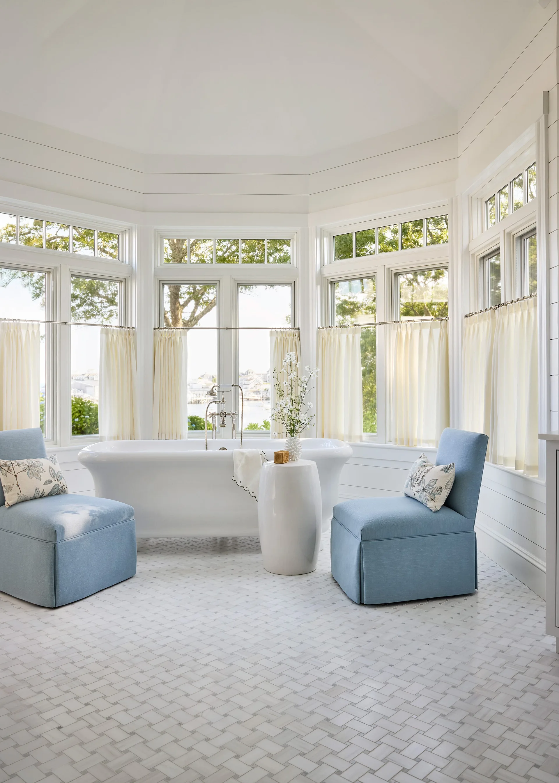 A white freestanding bathtub stands before a large bay window flanked by two light blue upholstered armchairs on a basketweave tile floor.