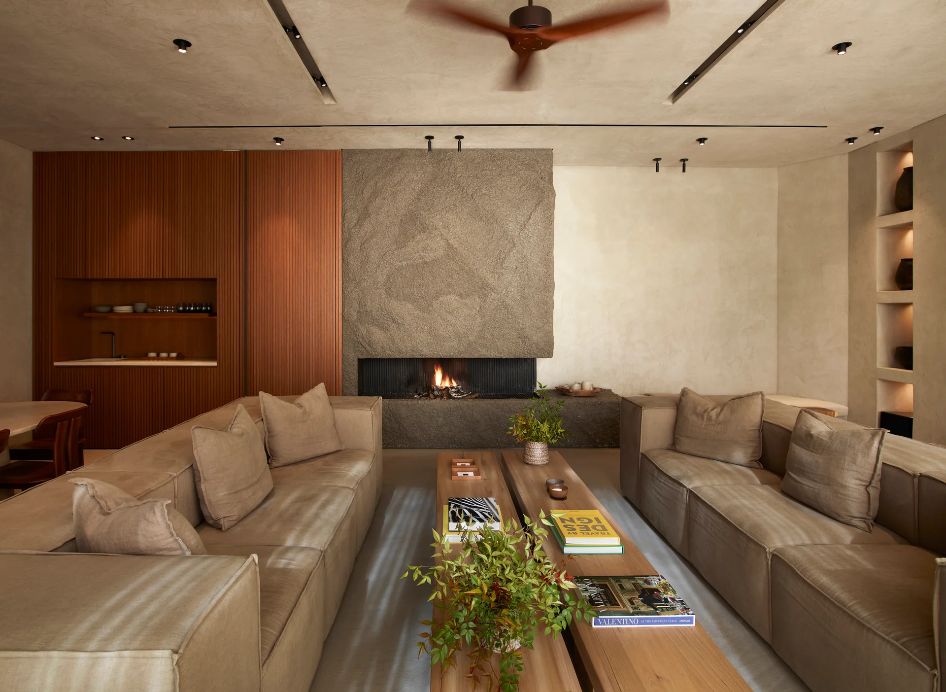 Two symmetrical beige sofas face each other across wooden coffee tables in a living room featuring a massive, rough-textured stone fireplace.