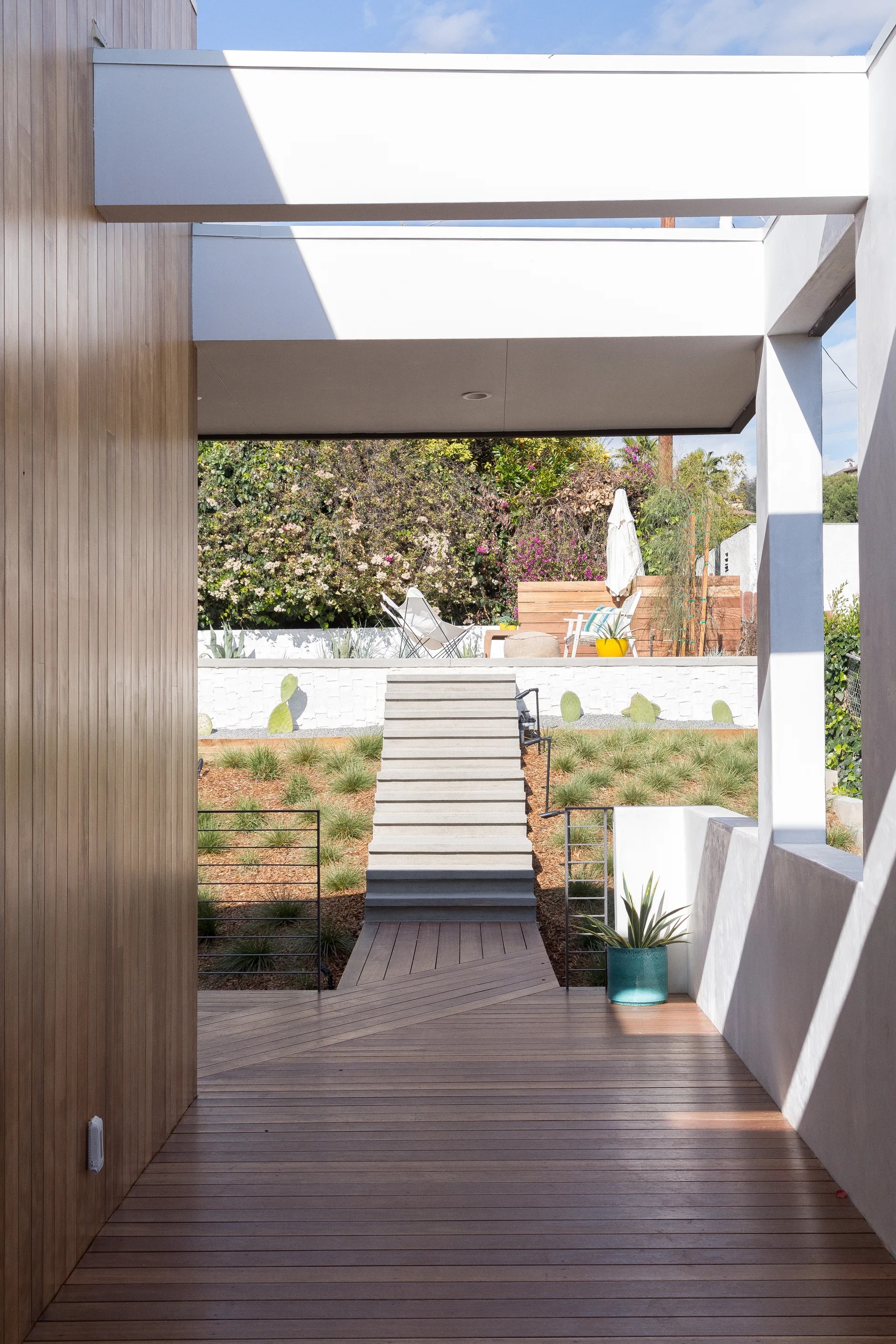 A view from a covered wooden deck looking down a short pathway toward concrete steps leading up to a terraced garden with lounge seating.