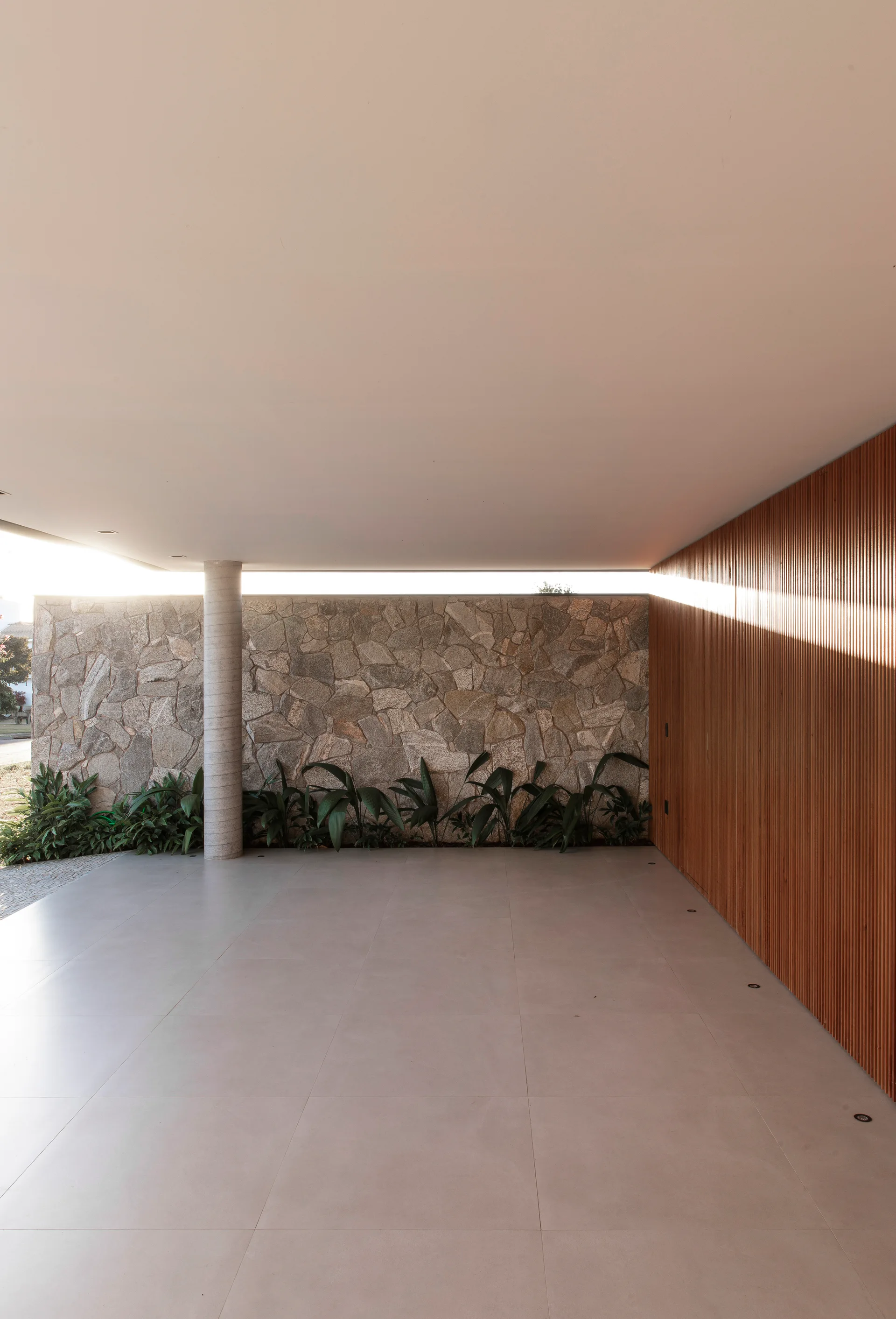 A minimalist covered patio features a vertical wood-slat wall, a rustic stone wall with green plants at the base, and large-format floor tiles.