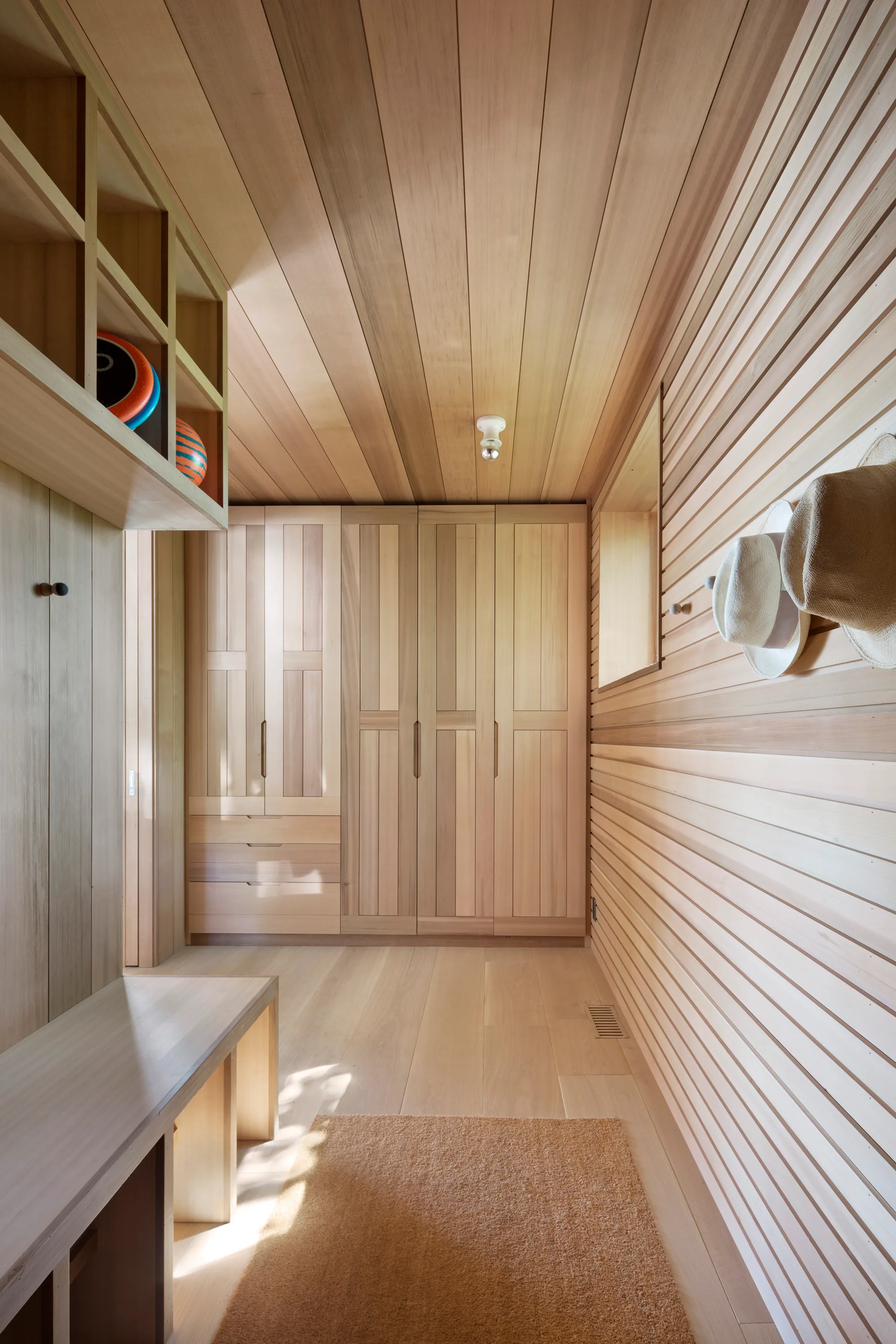 A modern mudroom features light wood paneling on the walls and ceiling, built-in wooden cabinets, a matching bench, and wall hooks with hanging hats.