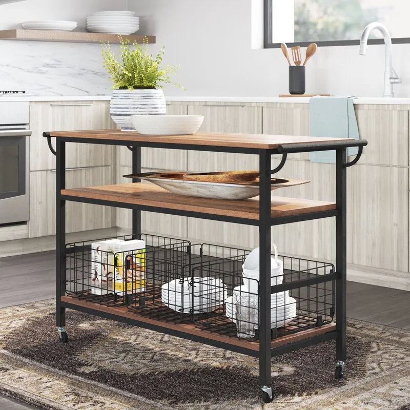 An industrial kitchen cart with black metal framing and wood shelves sits on a patterned rug, holding wire storage baskets and white dishes.