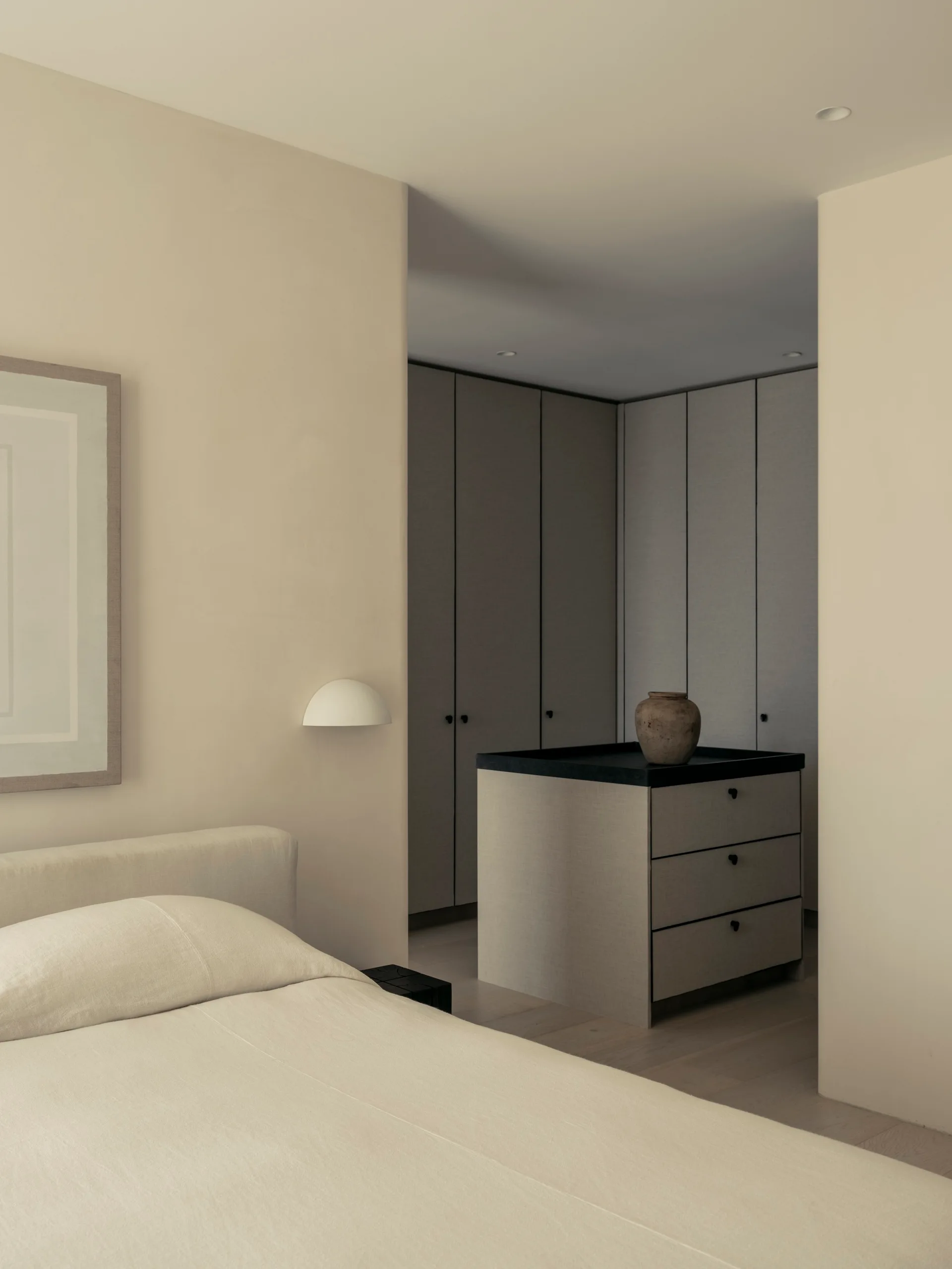 A beige bedroom features an upholstered bed and a wall sconce, looking into a walk-in closet with textured wardrobes and a dresser island.
