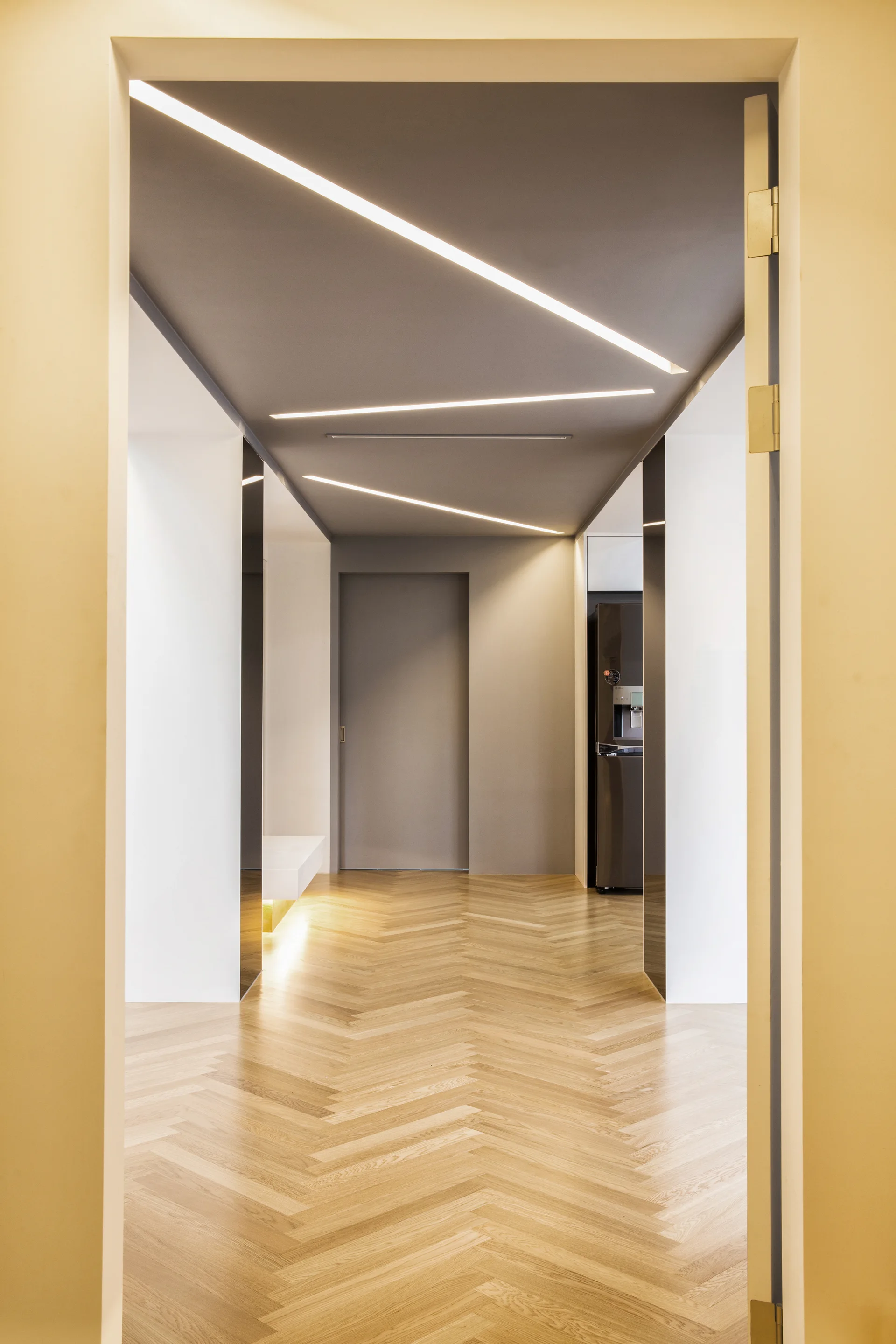 A modern interior hallway features light herringbone wood flooring, white walls, and a dark grey ceiling with angled recessed linear lights.