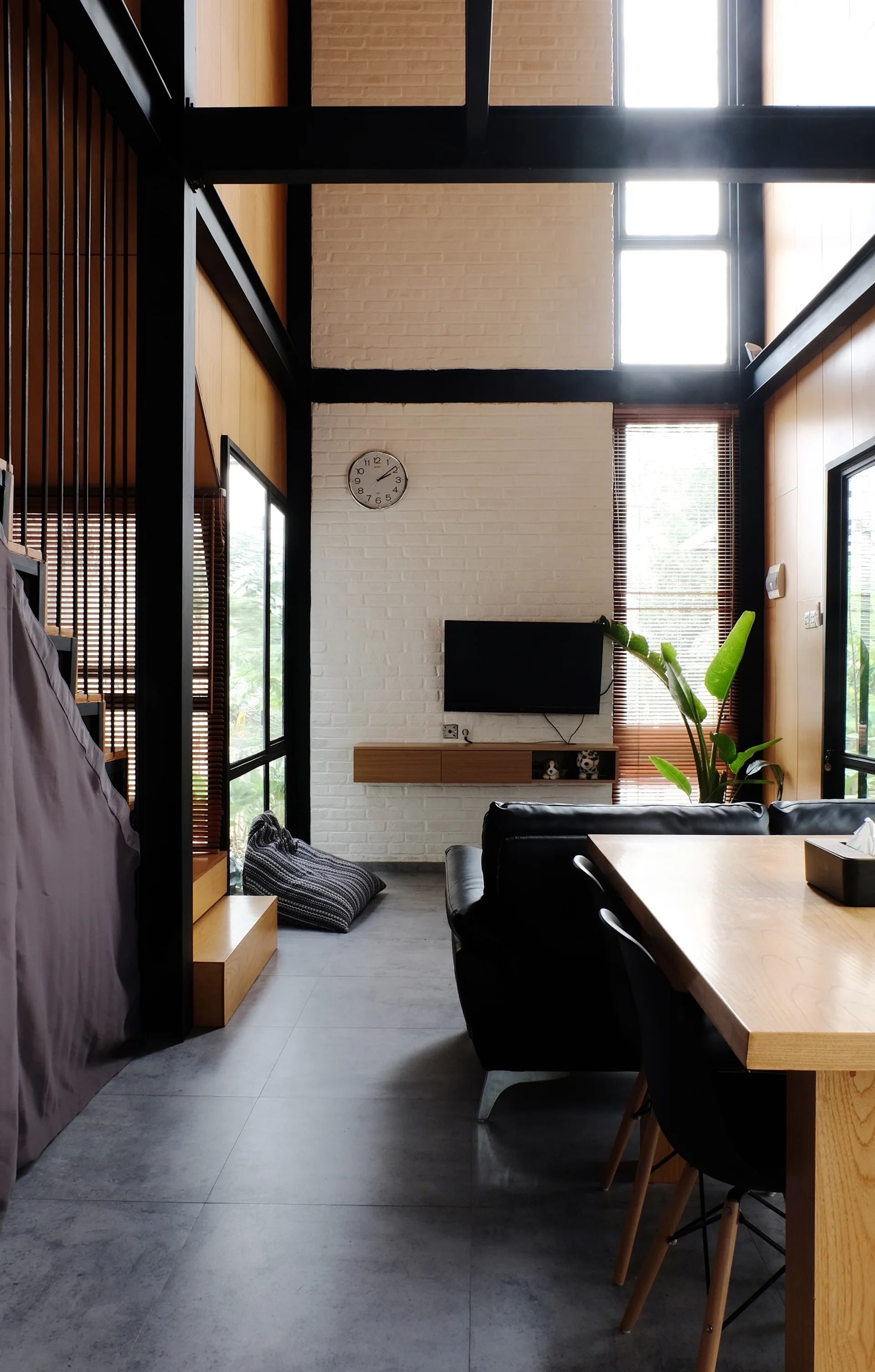 A double-height living room featuring a white painted brick media wall, a black leather sofa, a wooden dining table, and exposed black metal beams.