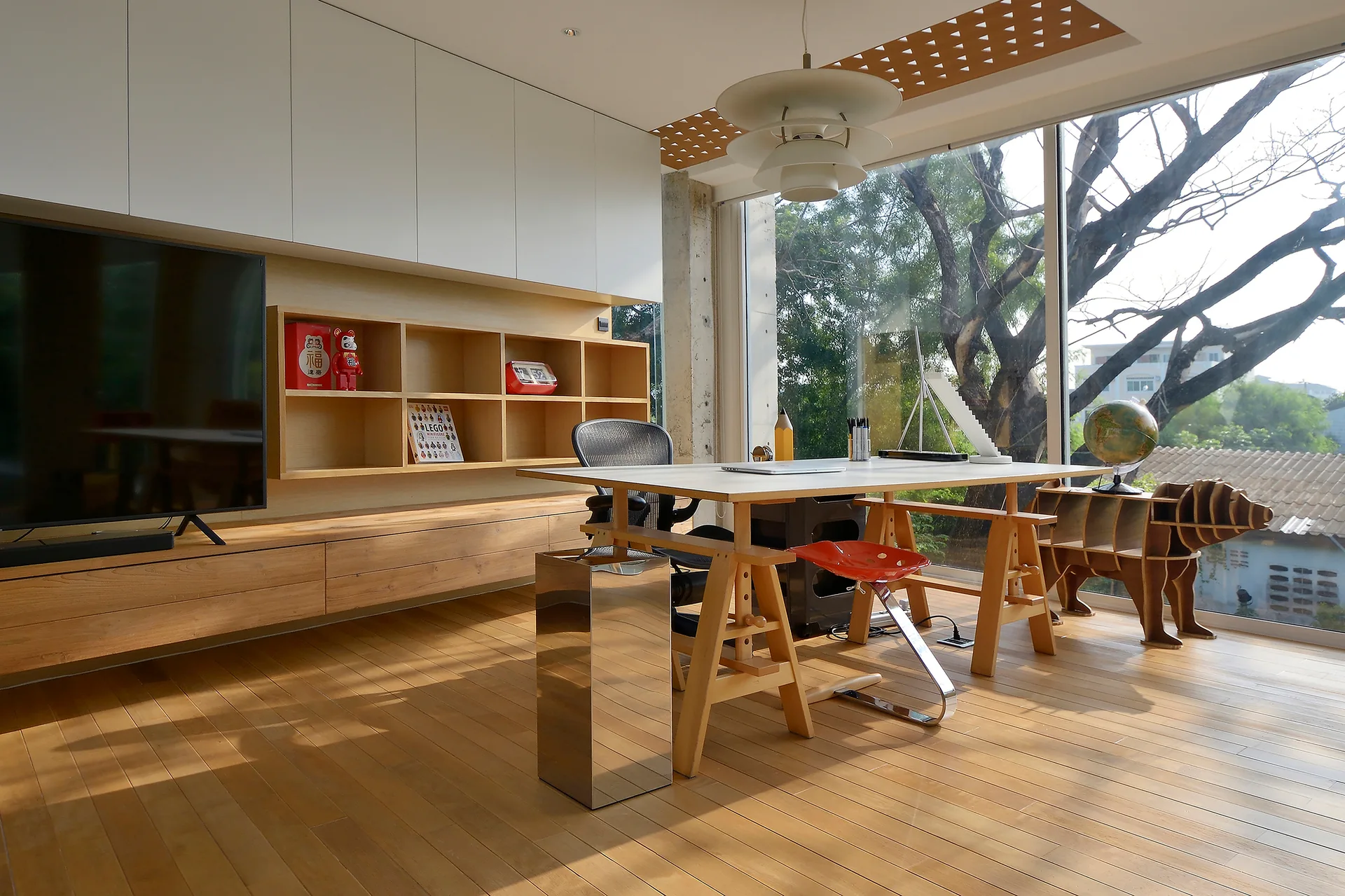 A bright minimalist home office features a wooden trestle desk, custom white and wood built-in shelving, a large flat-screen TV, and floor-to-ceiling windows.