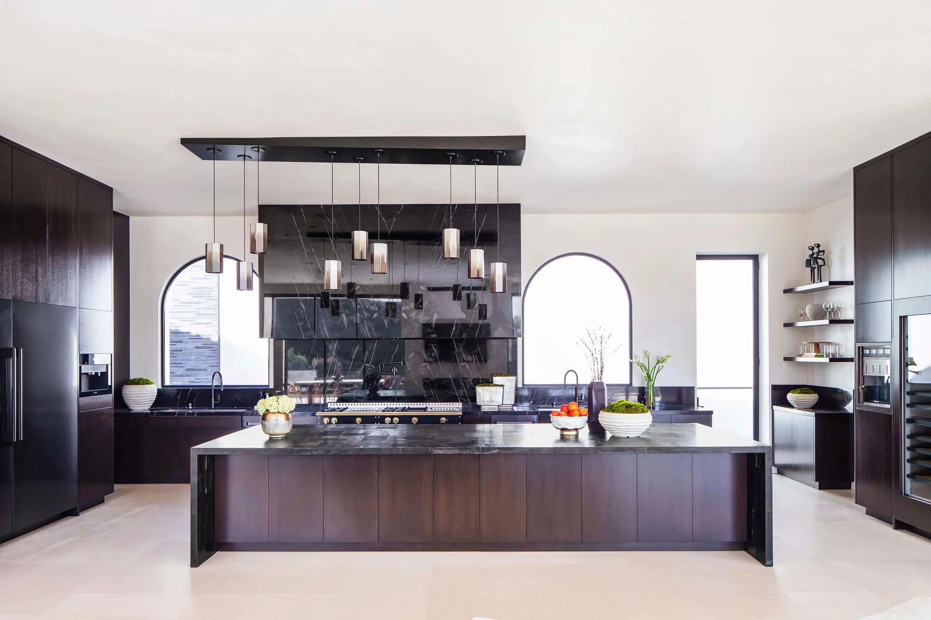 A modern kitchen featuring a dark wood island, matching cabinetry, arched windows, a black marble backsplash, and a cylindrical pendant light fixture.
