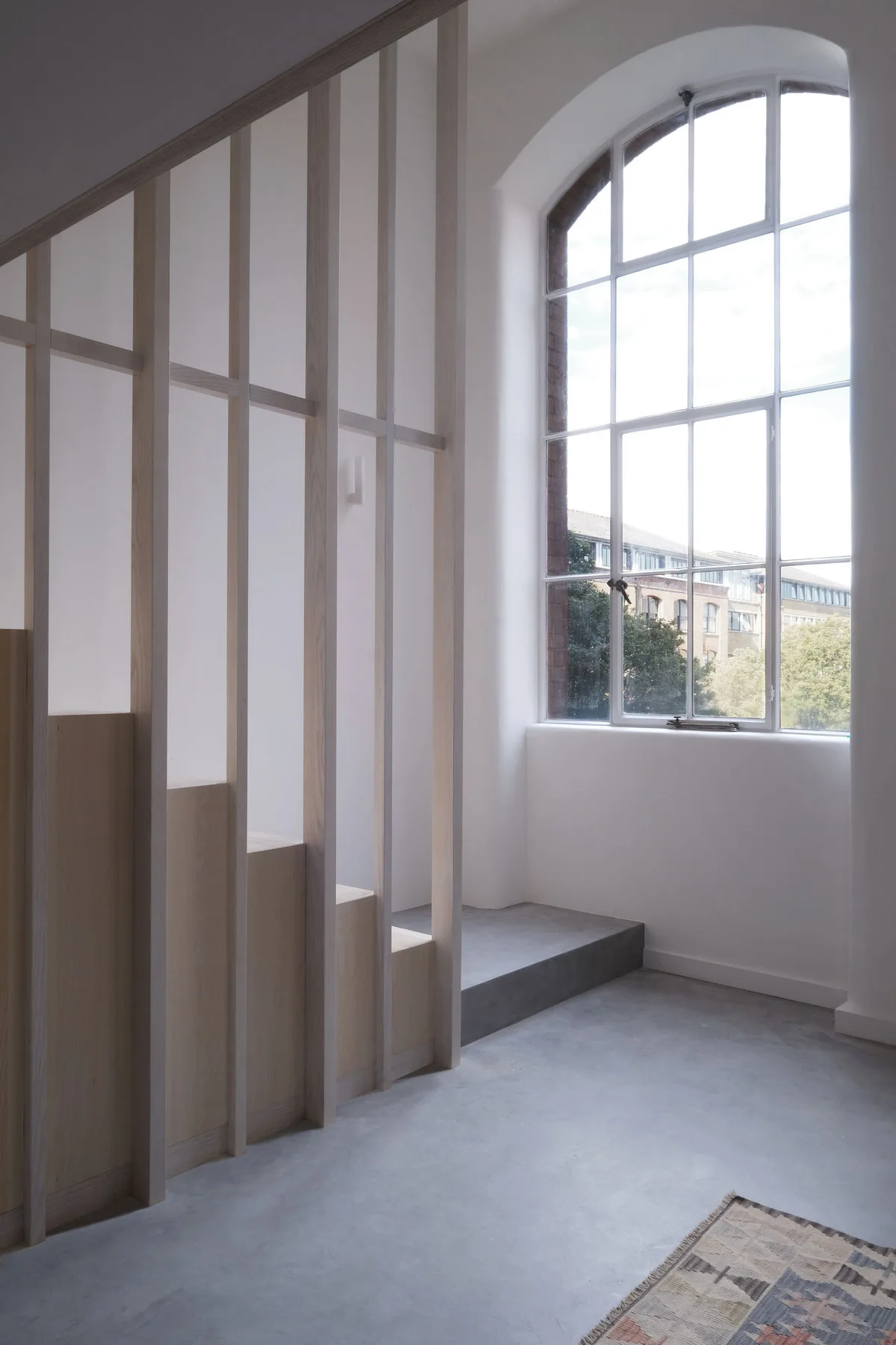 A minimalist interior features a light wood staircase with vertical slats next to a large arched window and grey concrete flooring.