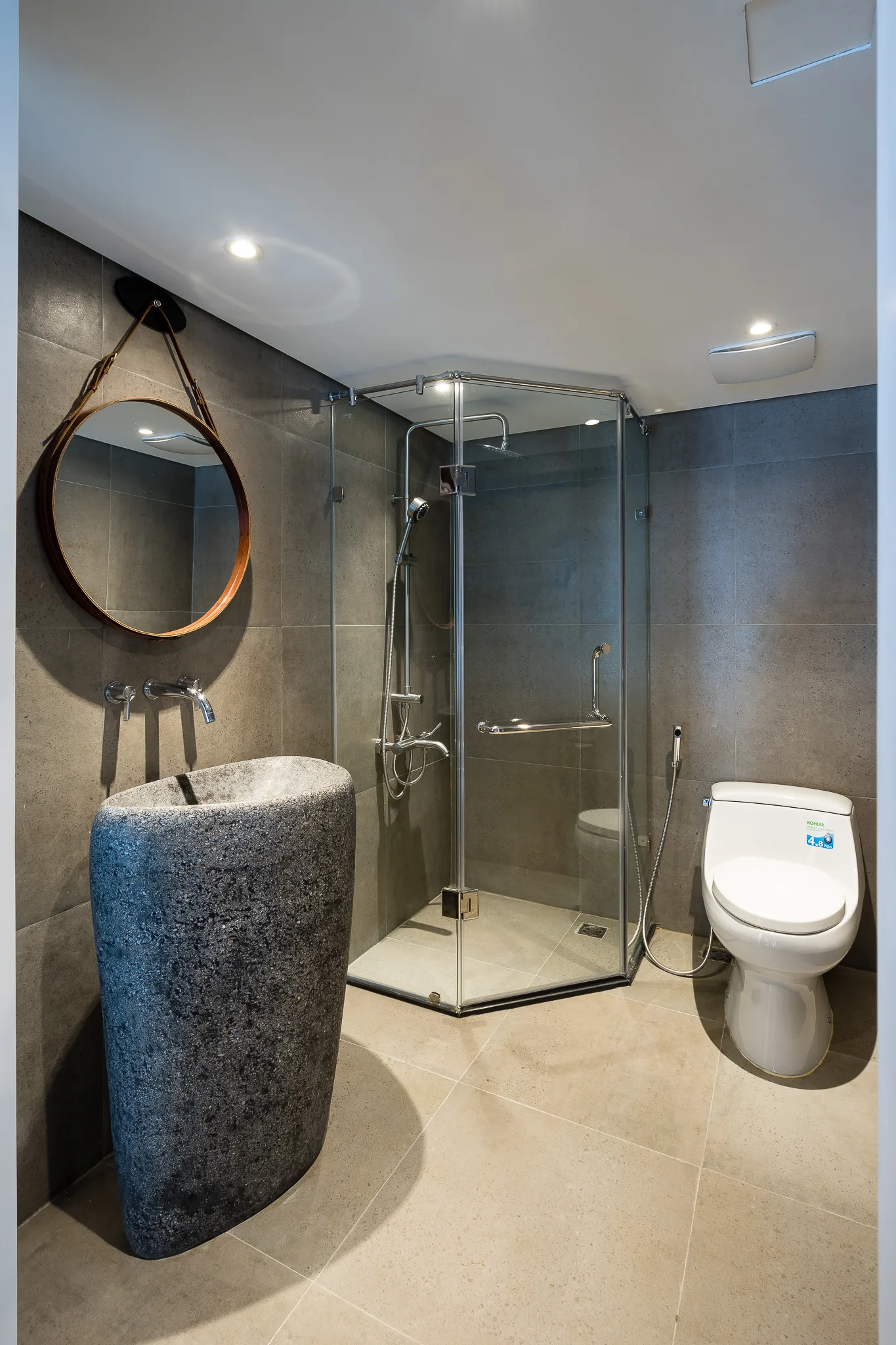 A modern bathroom features a dark carved stone pedestal sink, a round hanging mirror, a glass corner shower, and grey tiled walls.