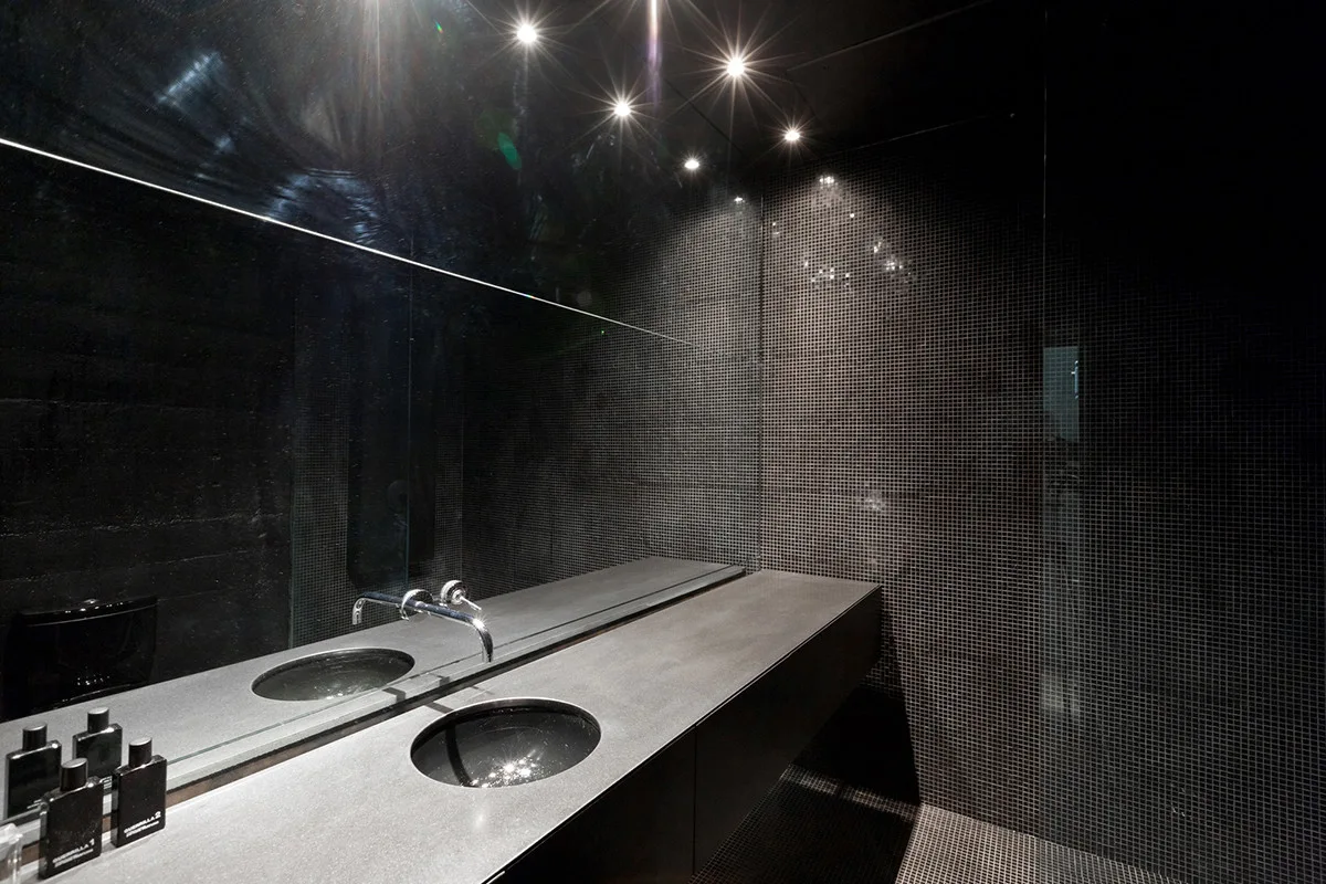 A minimalist bathroom features black mosaic tiles, a dark stone vanity with a chrome faucet, and a large mirror reflecting ceiling spotlights.