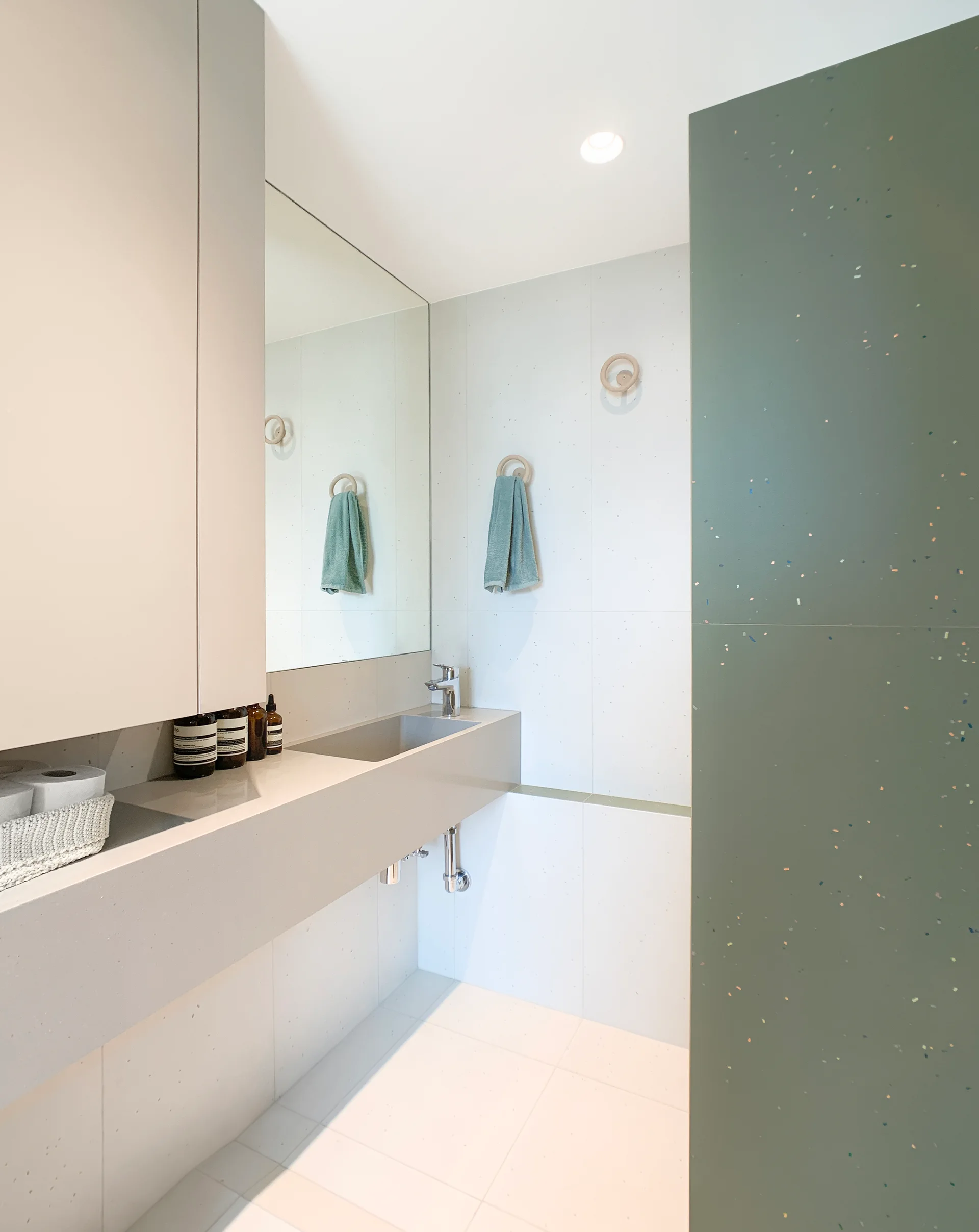 A modern minimalist bathroom featuring an integrated grey stone vanity sink, a large wall mirror, and a sage green terrazzo accent panel.