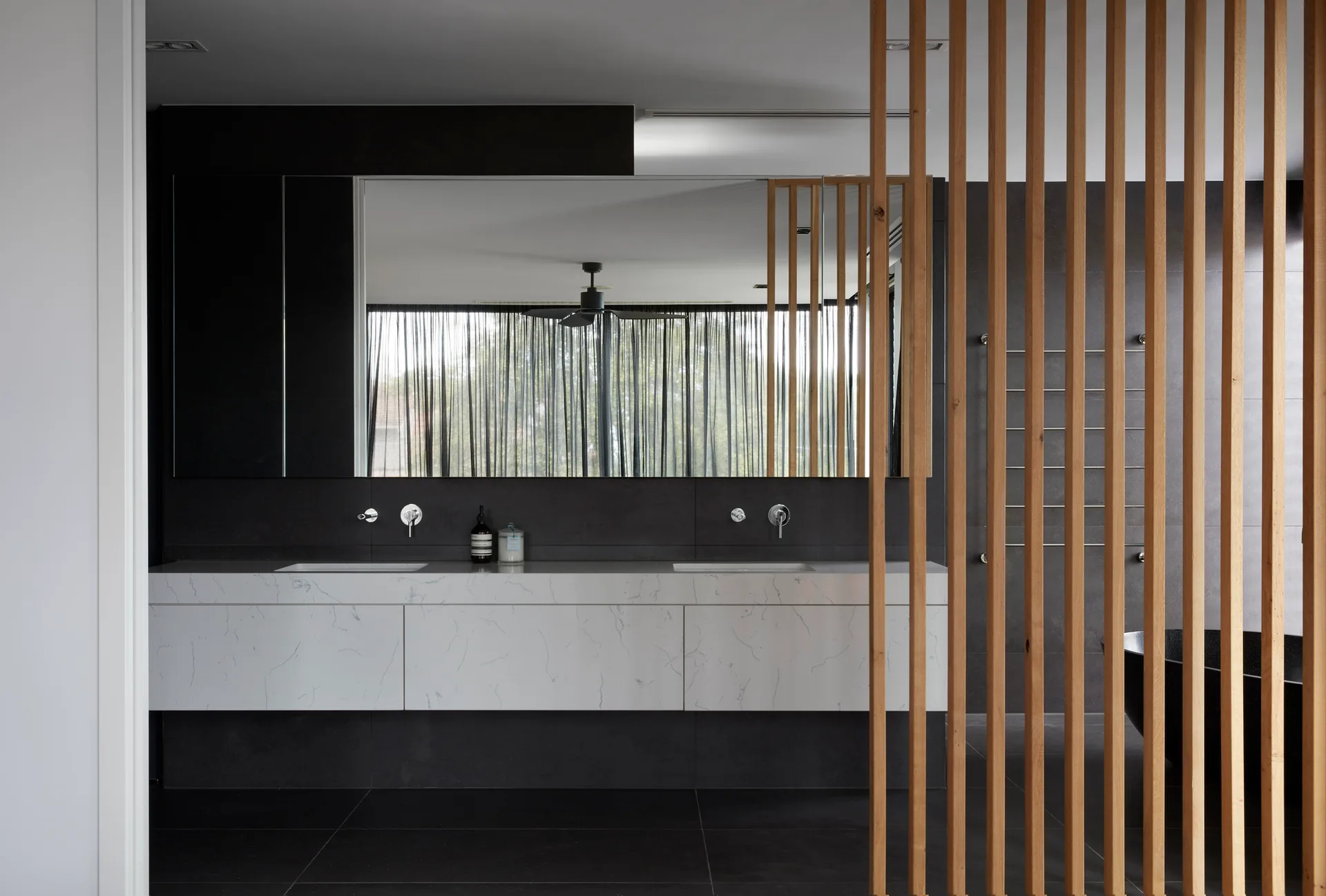 A modern bathroom features a white veined marble floating vanity, a large wall mirror, and a vertical wood slat room divider.