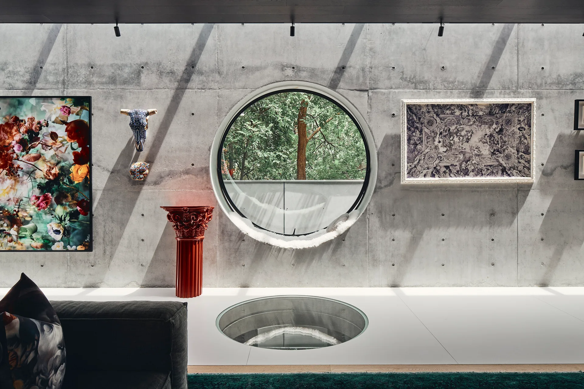 A modern interior space features an exposed concrete wall with a large circular window, a glass floor insert, and a decorative red column pedestal.