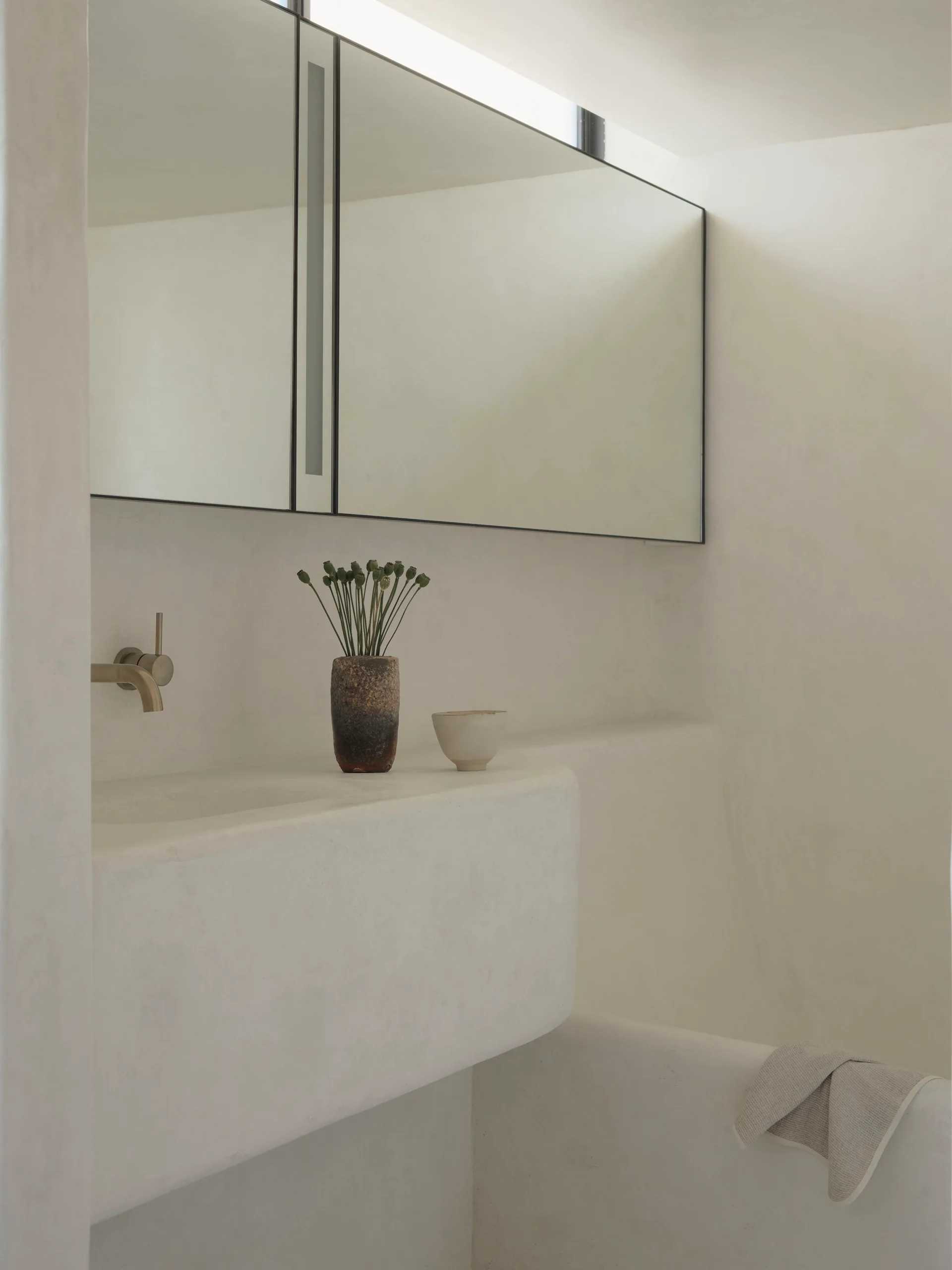 An off-white plaster bathroom features an integrated vanity sink with a bronze wall-mounted faucet under a large mirrored cabinet.