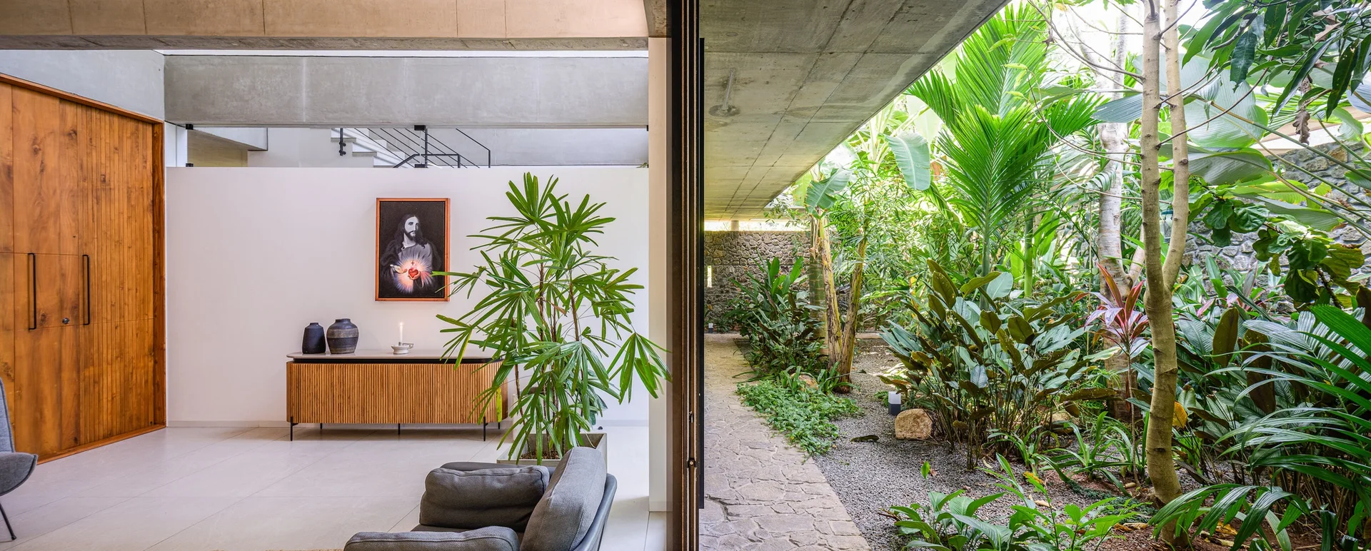 An indoor-outdoor living room with a slatted wood sideboard, an exposed concrete ceiling, and a wide doorway opening to a lush tropical courtyard garden.