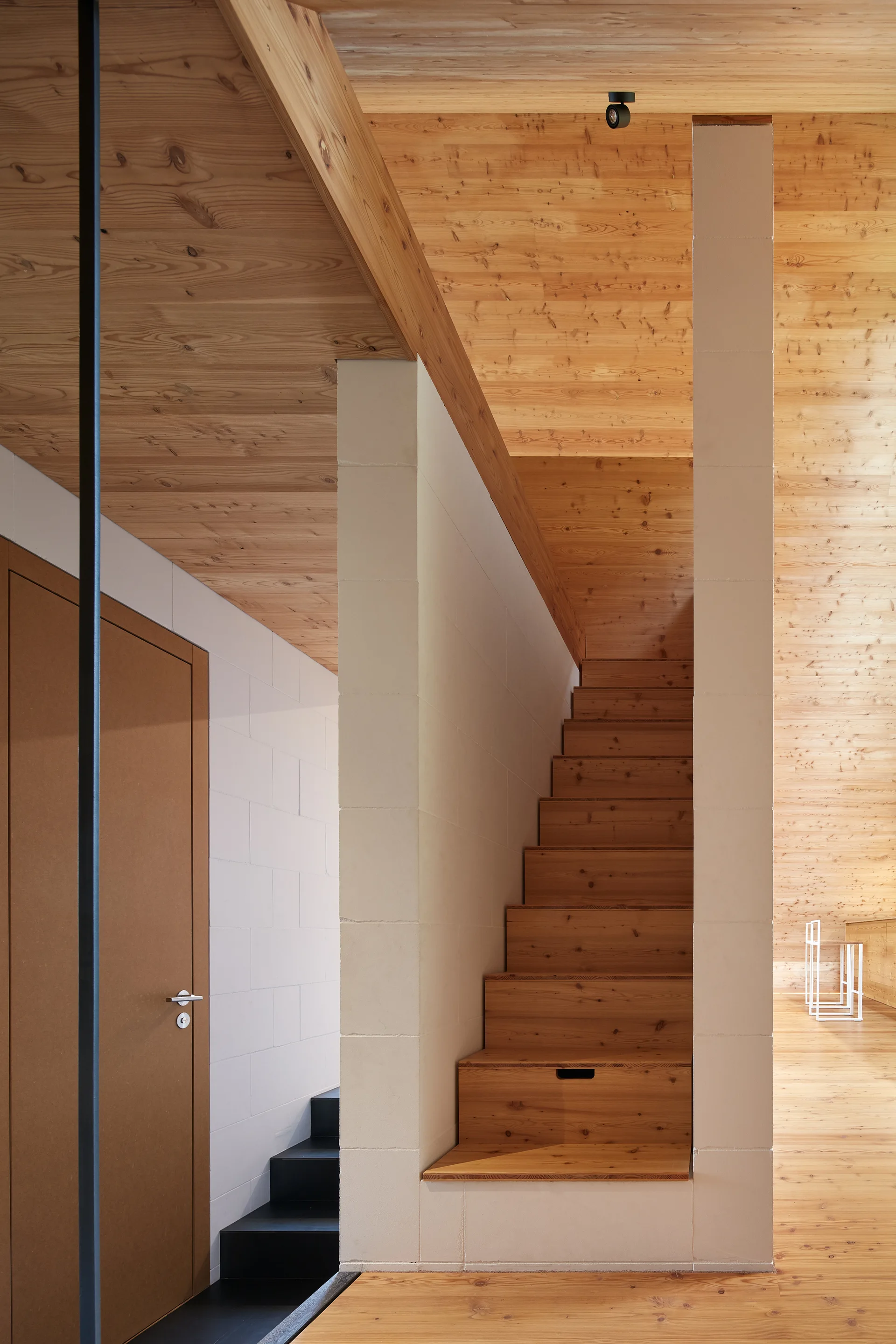 A natural wood staircase with storage built into the bottom step is framed by white concrete block columns under a sloped wooden ceiling.