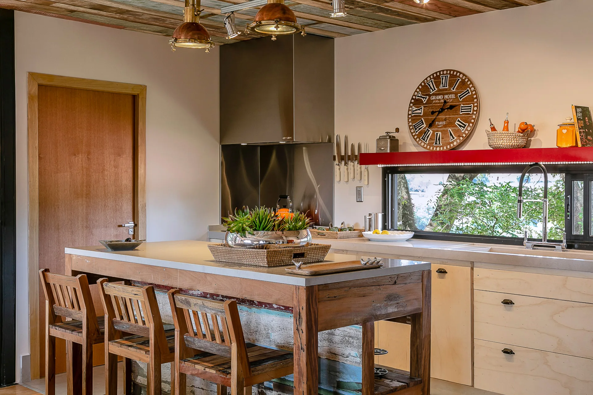 A rustic kitchen features a wooden island with bar stools, light wood cabinets, and a long horizontal window behind the sink counter.