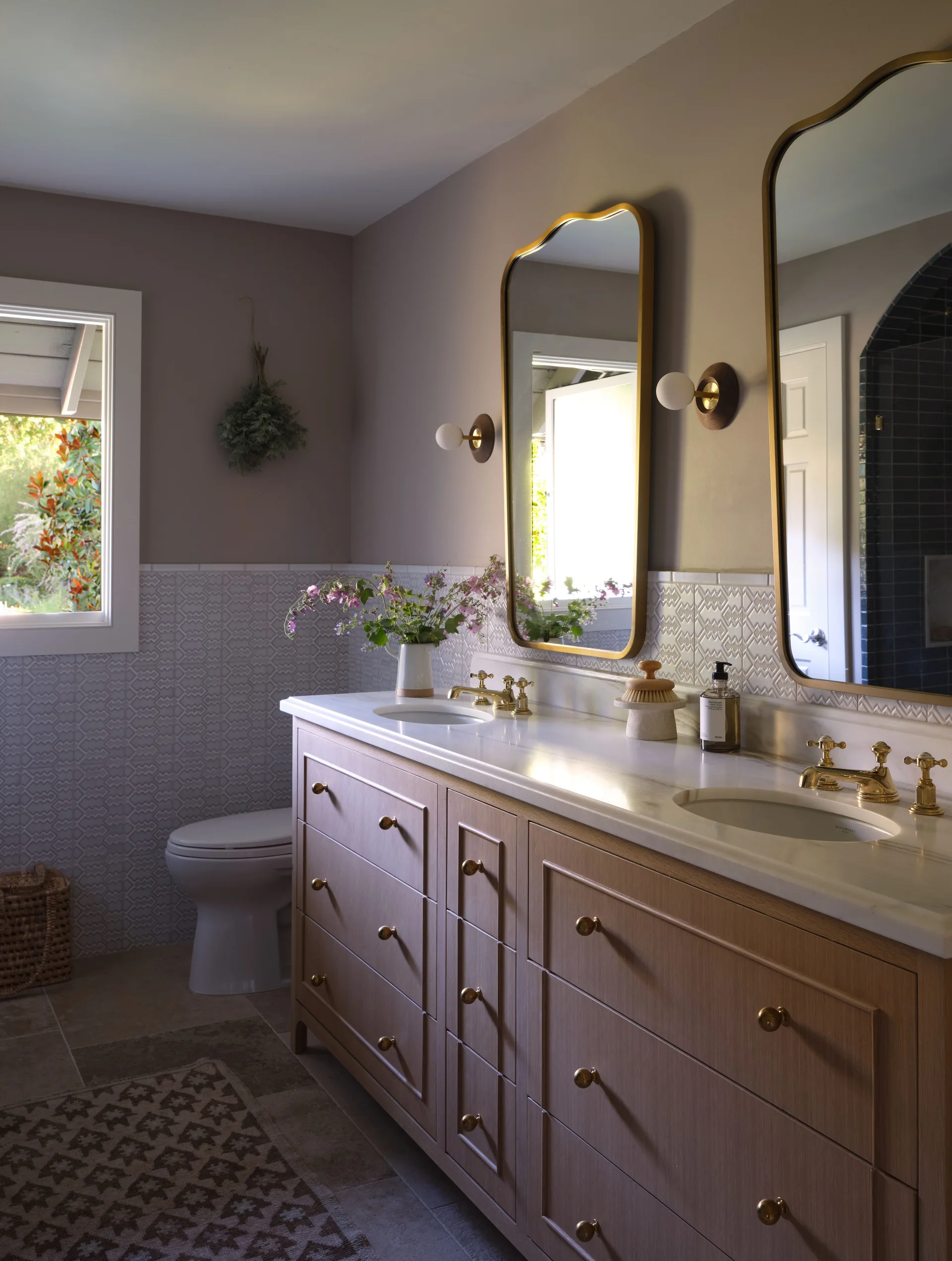 A light wood bathroom vanity with a white marble top, gold faucets, dual gold-framed mirrors, and geometric wainscot tile on taupe walls.