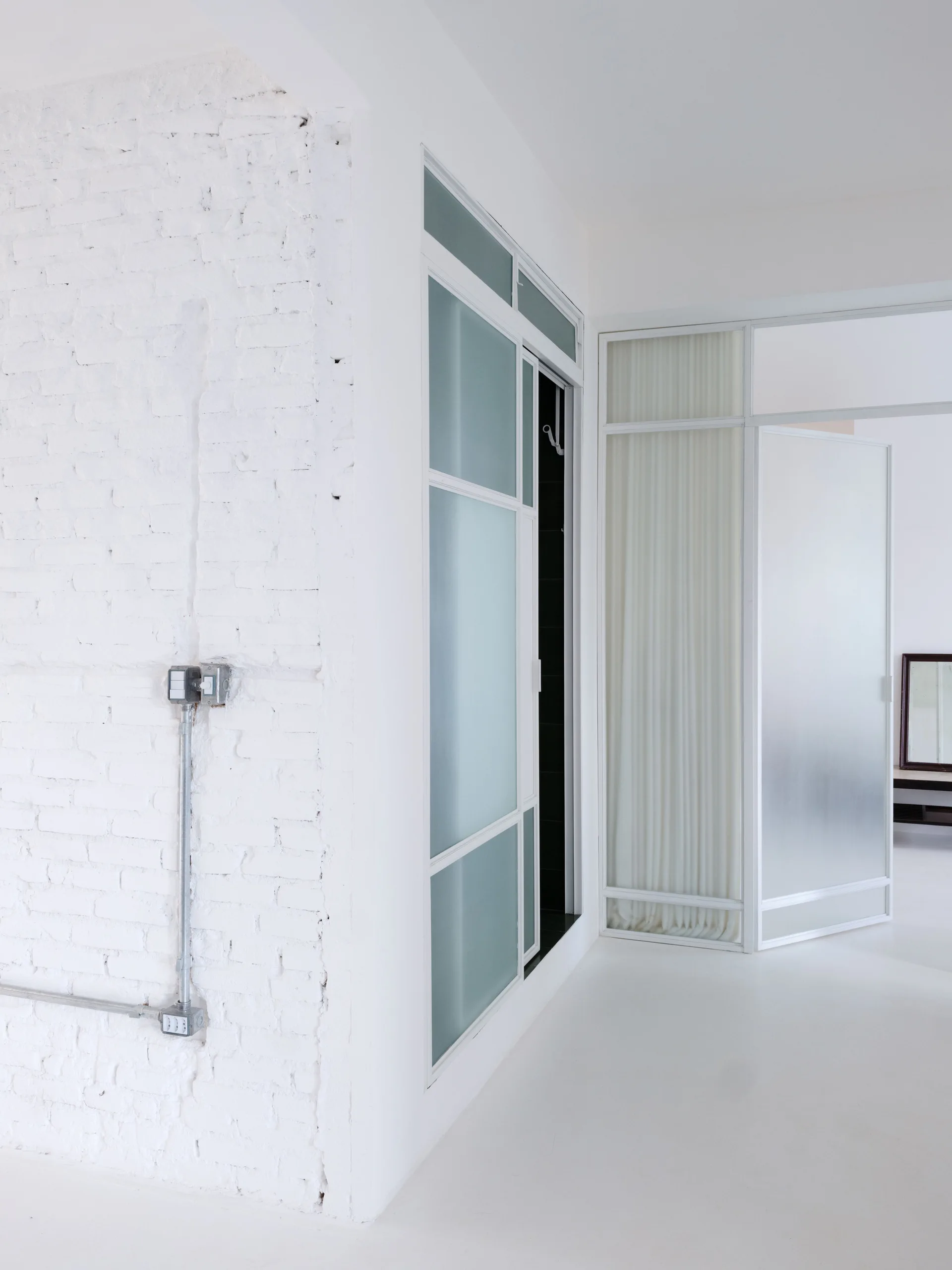 A white painted brick wall with exposed metal conduit stands next to a metal-framed room divider with frosted glass panels.