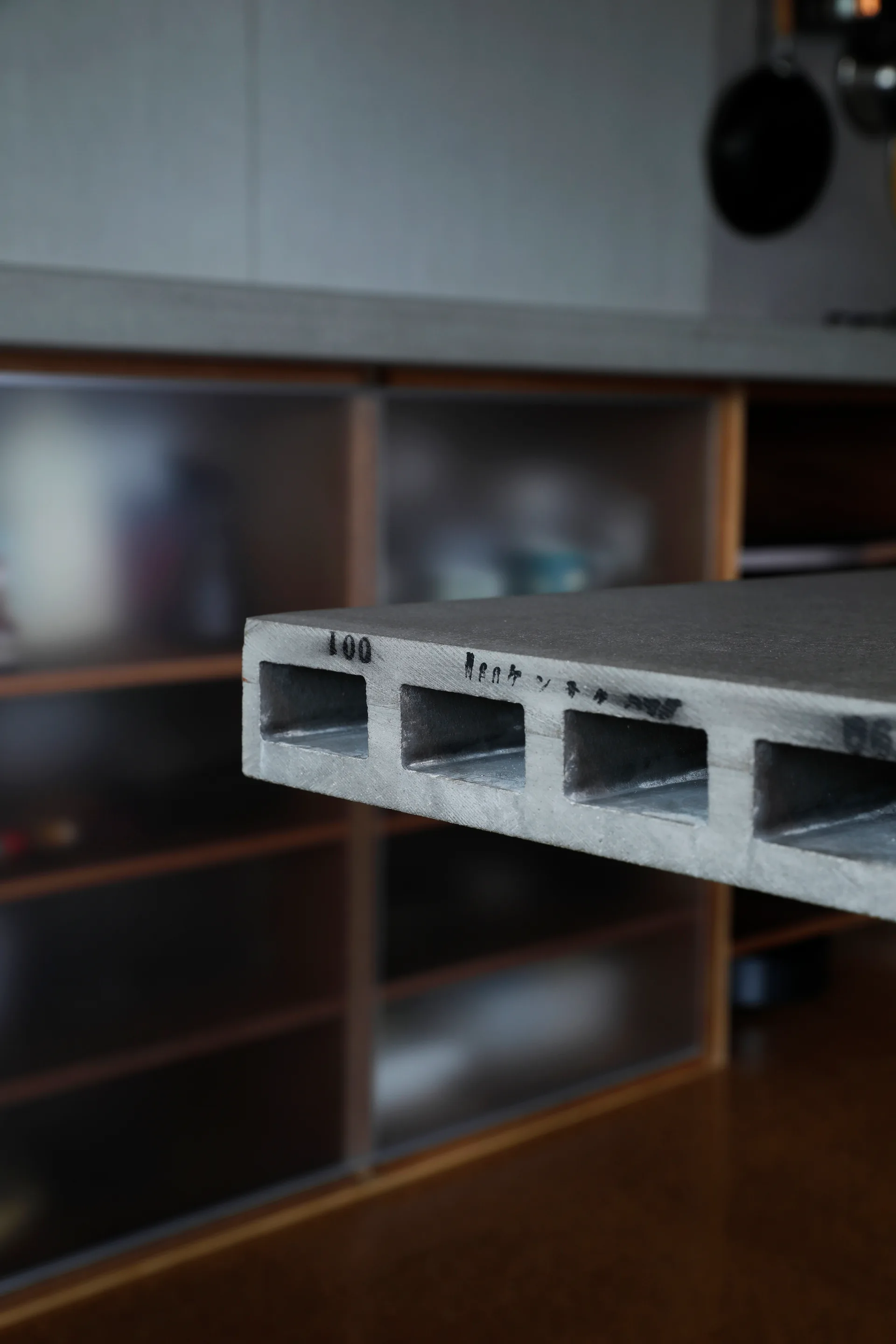 A close-up of a grey hollow-core concrete panel sample showcasing rectangular channels on the edge in front of blurred kitchen cabinets.