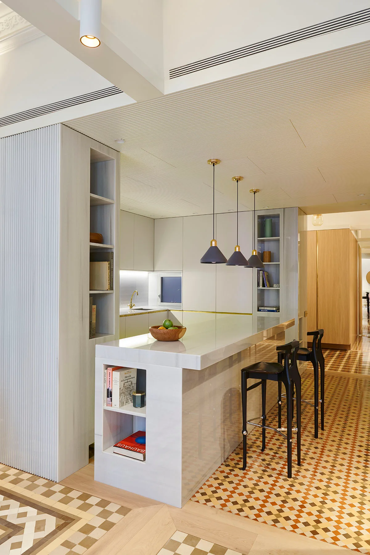 A modern kitchen featuring a white stone island with built-in bookshelves, black bar stools, dark pendant lights, and geometric patterned floor tiles.