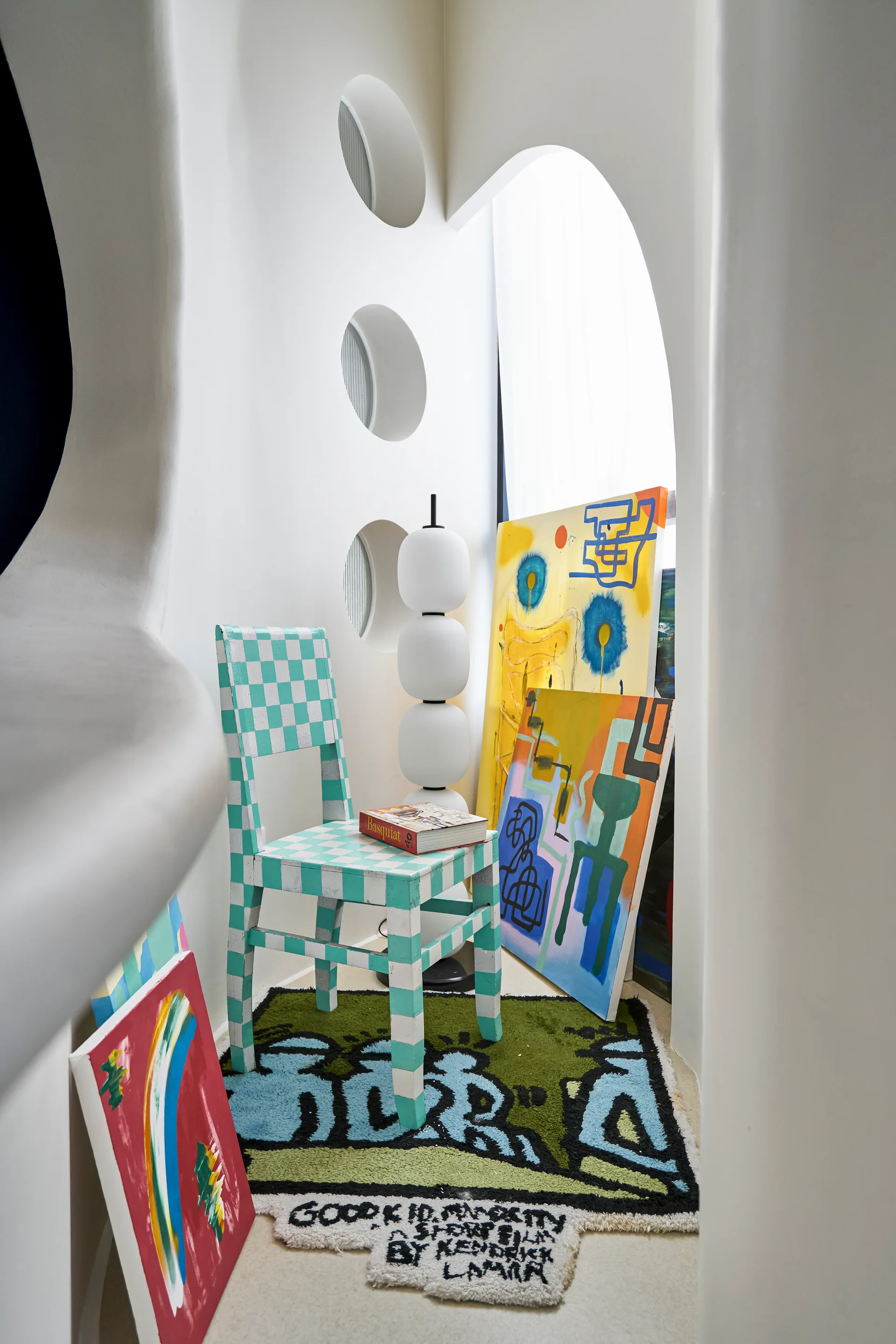 A small white plaster alcove features a green and white checkered chair, a stacked white orb floor lamp, and colorful abstract paintings.
